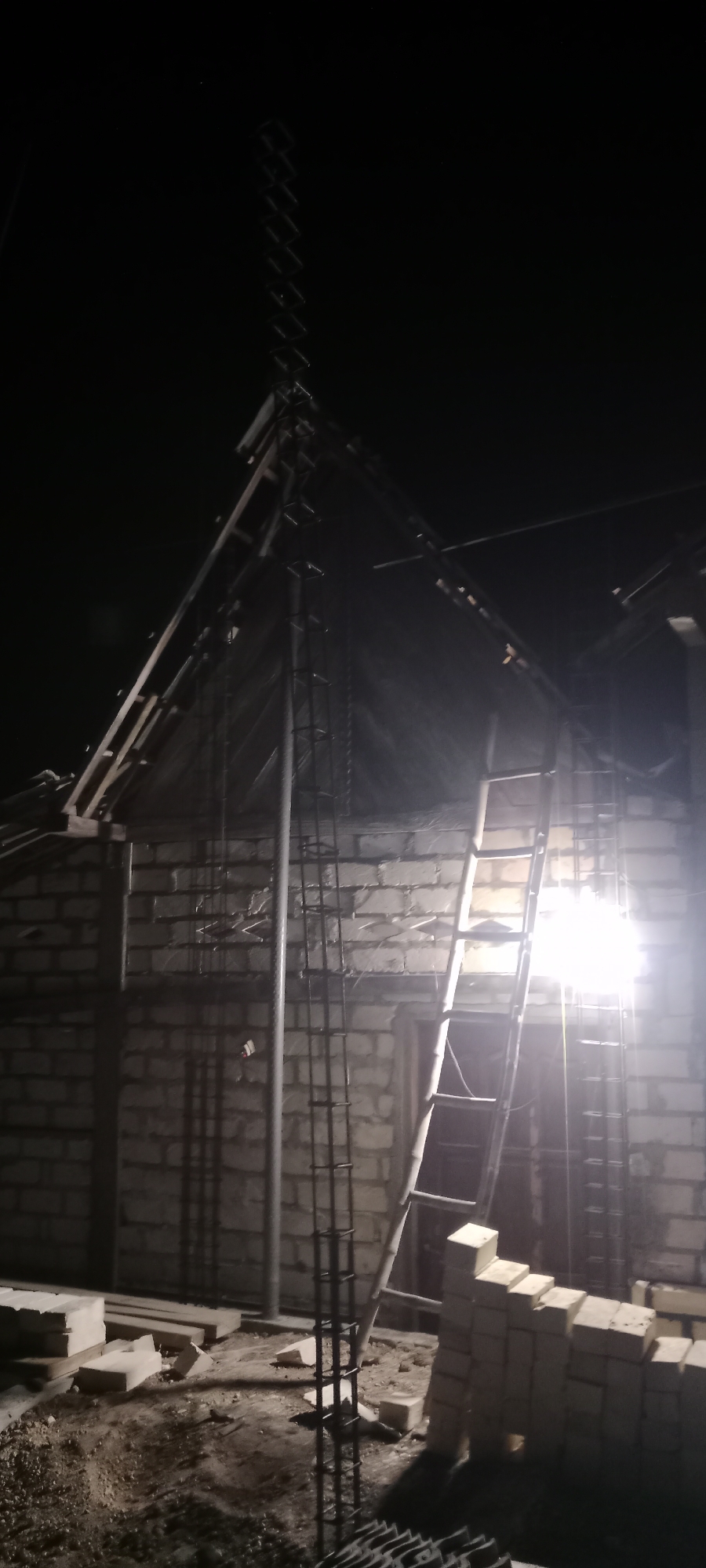 An outdoor, nighttime scene depicts a construction site in Soko, Indonesia. A building is under construction, with its brick walls partially completed. The roof structure, made of wood and metal beams, is exposed. A tall, vertical metal rebar cage extends upwards from the ground, reaching into the partially constructed roof. A wooden ladder leans against the building, providing access to the upper levels. Piles of bricks are stacked near the building. The ground is dirt and debris. A bright light source, possibly a construction lamp, illuminates a section of the building and the surrounding area, creating strong shadows. The overall impression is that of ongoing construction work at night.