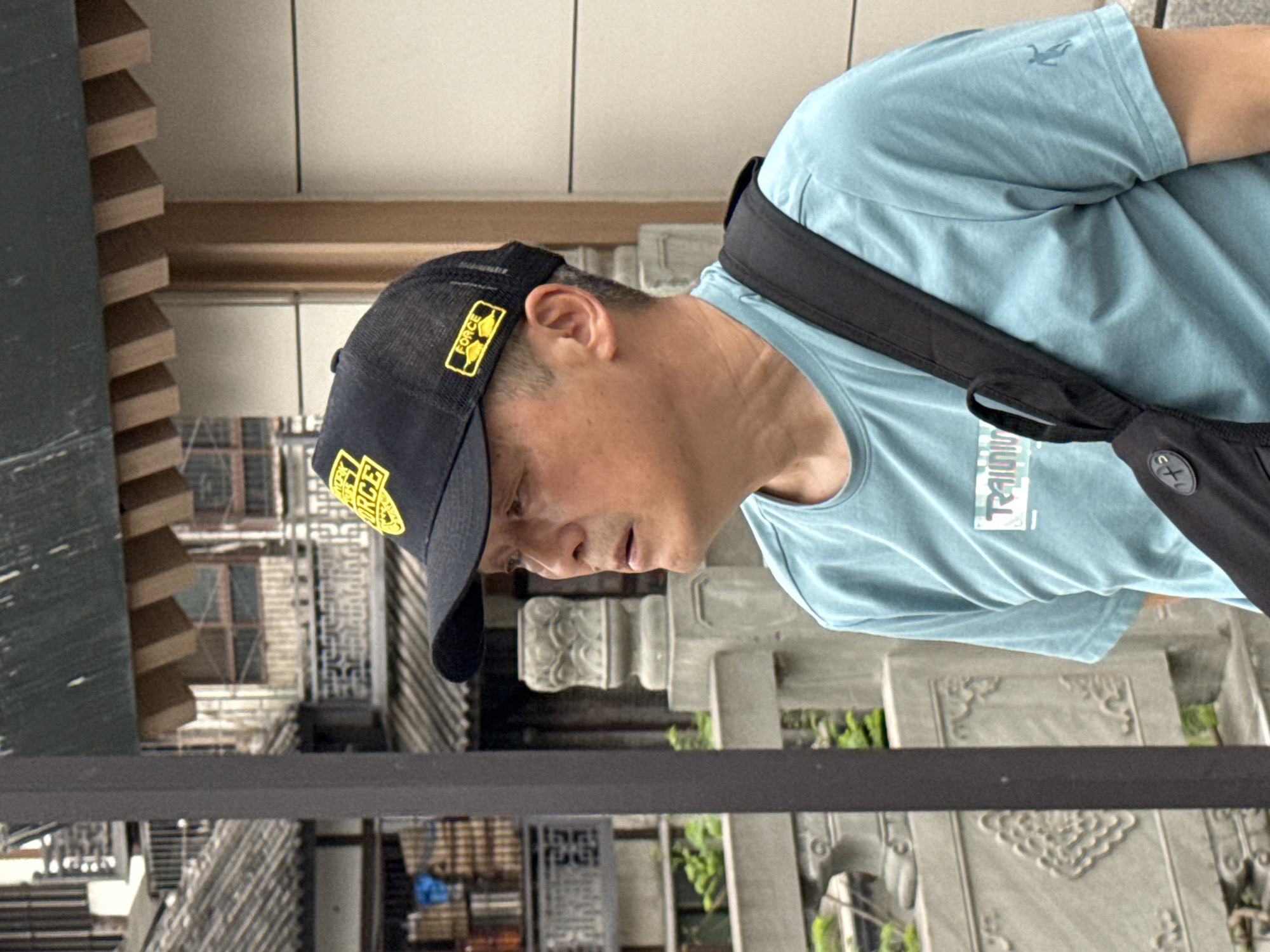A middle-aged Asian man wearing a black baseball cap, light blue t-shirt, and a black shoulder bag is looking down at an ornate stone fence, which is in front of a traditional Chinese building. The location appears to be in the Shapingba District, China.