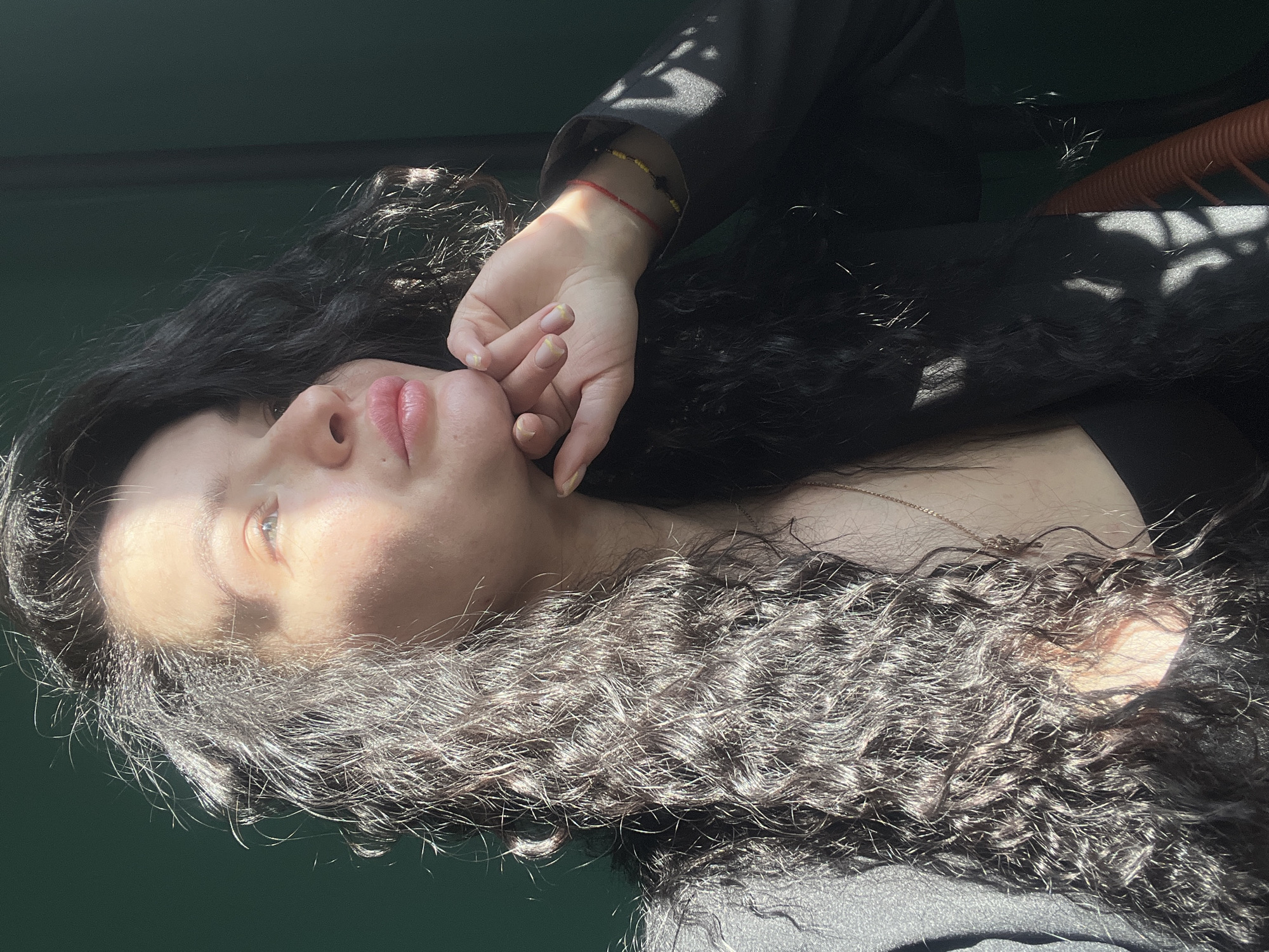 A medium, eye-level shot shows a woman with long, dark curly hair. Her head is turned slightly upwards and her hand is touching her chin. She is wearing a black garment. A red and yellow beaded bracelet is visible on her wrist. The lighting is from above, casting shadows across the scene. The background is dark and indistinct. The location is unknown.