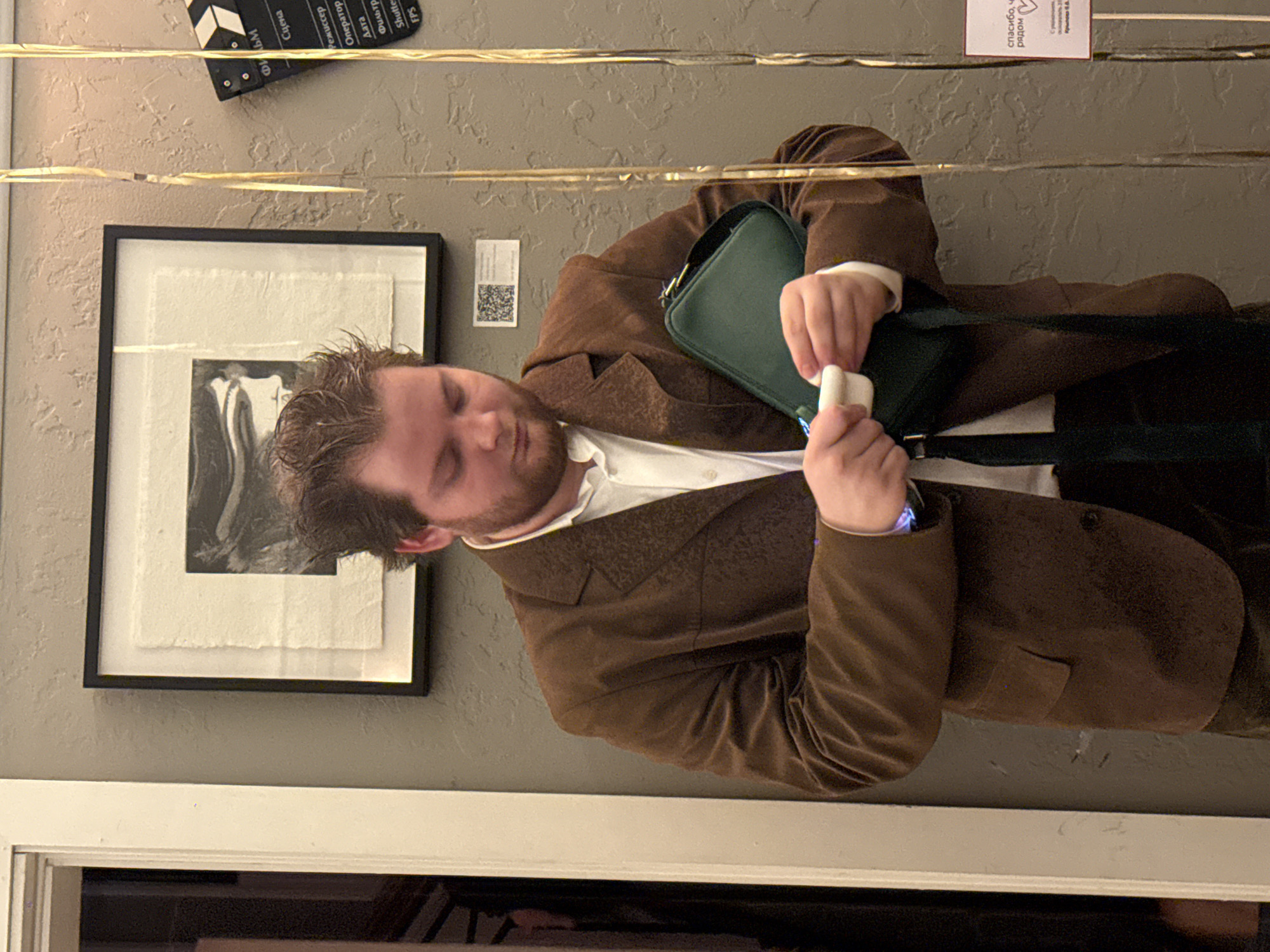 A man in a brown suit and white collared shirt is holding white earbuds in his hands. He is standing in front of a gray wall with a framed black and white picture. There is a green purse hanging on his shoulder. A movie clapboard is above the picture. A white sign is next to the picture with a QR code and Russian text on it. Gold tinsel hangs in front of the wall. The location is likely in Russia given the movie clapboard is written in Russian as well as the text on the white sign. Given the fact that a clapperboard is shown with information written in Russian, it can be assumed that this photo was taken in Moscow, Russia.
