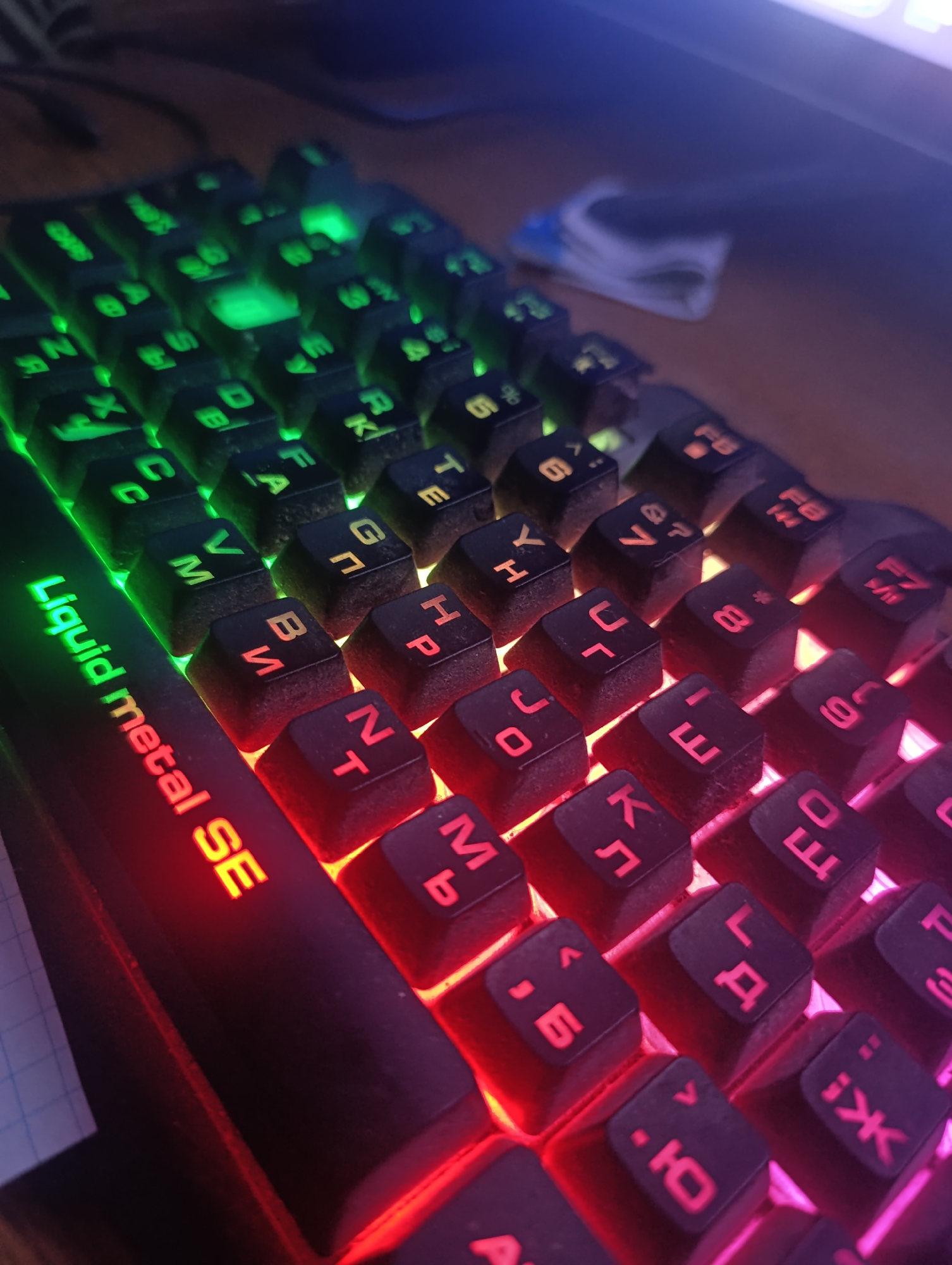 The image shows a close-up view of a black mechanical keyboard with backlit keys. The lighting is predominantly red and green, with some keys illuminated in pink and purple hues. The letters on the keys are in a contrasting color, with some appearing to be Cyrillic and others Latin. The brand name "Liquid metal SE" is visible in bright red letters along the left side of the keyboard. The keyboard is placed on a wooden surface, and a partially visible piece of paper with a grid pattern is to the left. The background is dark and out of focus, suggesting an indoor setting, possibly a desk. The image does not contain any people, and there is no discernible information indicating a specific location in the city of Zaliznyi, Russia.