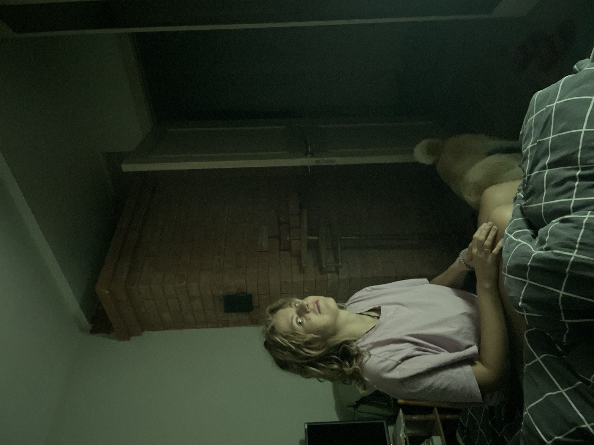 The image shows a dimly lit interior scene. A woman with blonde curly hair, wearing a light purple t-shirt and a bracelet, is sitting on a bed, partially covered with a dark gray and white plaid blanket. Beside her, on the bed, sits a tan-colored dog. Behind the woman and the dog is a large brick fireplace. Above the fireplace is a dark, recessed ceiling.
The location is not possible to determine from the image. Russia and Sestroretsk cannot be determined from the visual data.