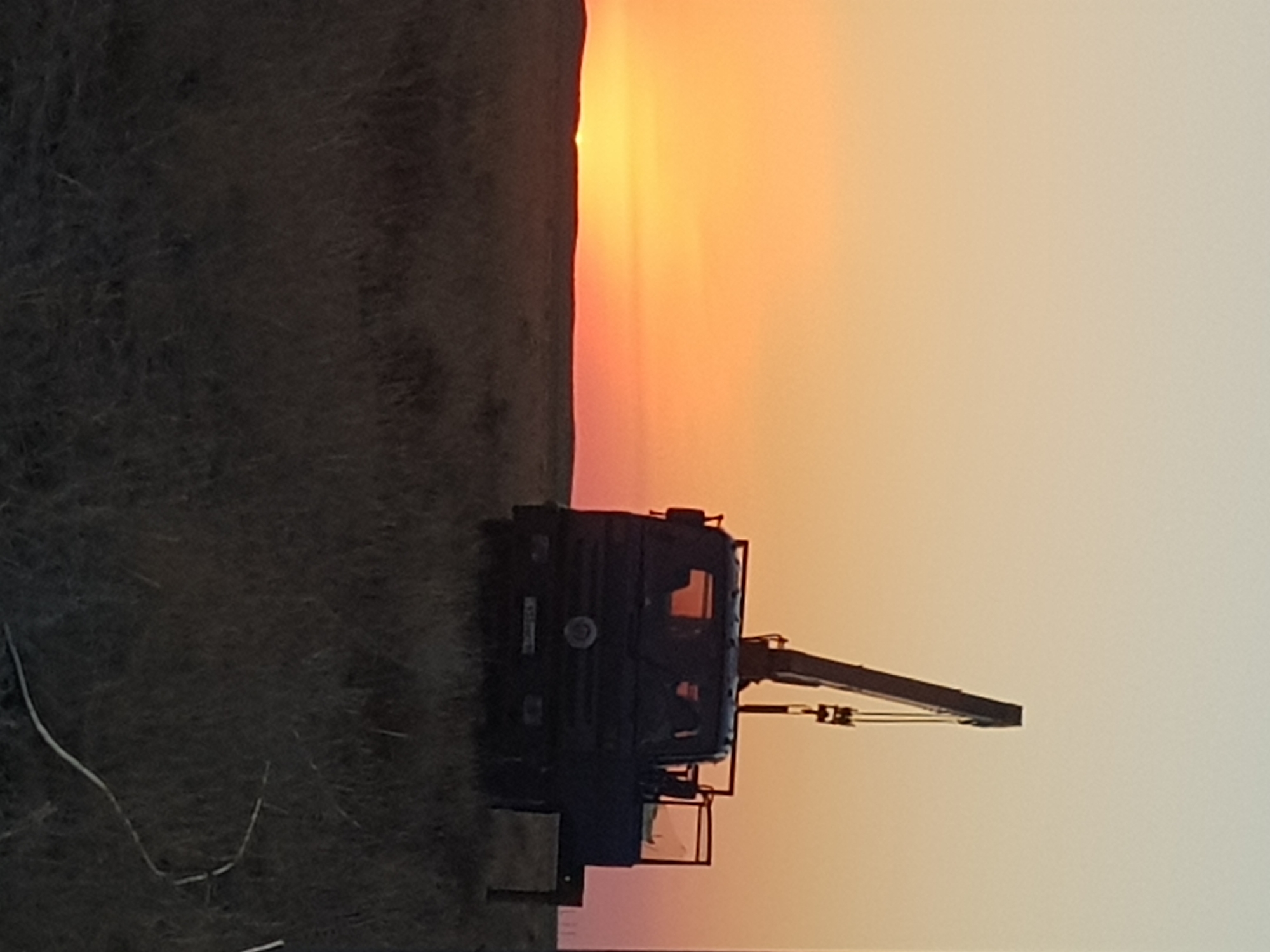 A dark truck with a crane attached is parked in a grassy field at sunset. The sky is a gradient of orange and yellow, with the sun low on the horizon. The truck is silhouetted against the bright sky, and its crane is extended upwards. The field is covered in dry grass, and there are a few small hills in the distance. The scene appears to be in an undeveloped area, possibly a rural or remote location. The image was taken in the territory of the Semipalatinsk Test Site, Kazakhstan.