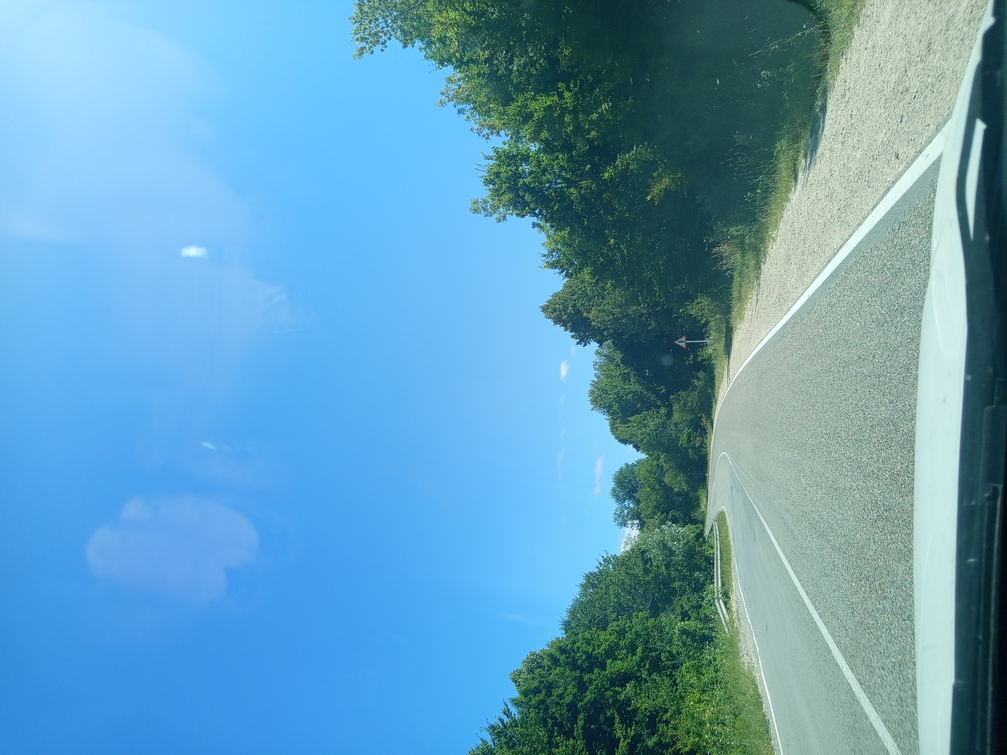 This is a point-of-view photograph taken from a vehicle traveling on a paved road through a wooded area, likely in or near Giaginskaya, Russia, given the context. The road curves to the right and is bordered by a gravel shoulder and dense green trees on both sides. A white line marks the edge of the road on the right, and a broken white line divides the lanes. A triangular warning sign is visible on the left side of the road in the distance. The sky is a clear, bright blue with some light, diffuse reflections, possibly from the windshield. The scene depicts a typical rural or highway drive on a sunny day.