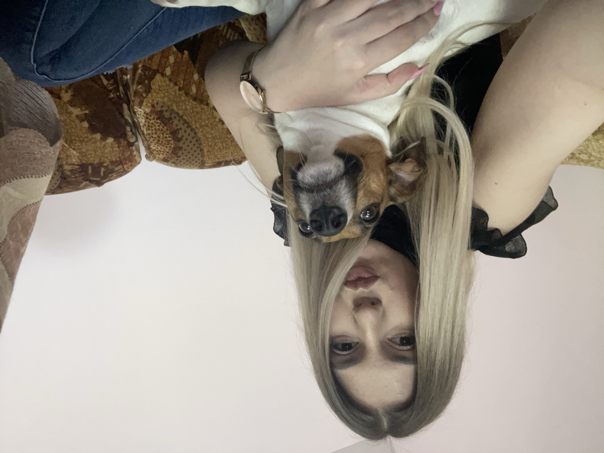 A selfie-style photograph captures a woman holding a dog, both positioned upside down relative to the frame. The woman has light blonde hair with darker roots, light skin, and dark eyes. She wears a black top, and a watch is visible on her left wrist. The dog, held in her arms, is predominantly white with brown and black markings on its head. They are situated on a sofa with a brown patterned upholstery, with the backdrop being a light pink wall. There is no information present in the image to specify the location as Burgas, Bulgaria.