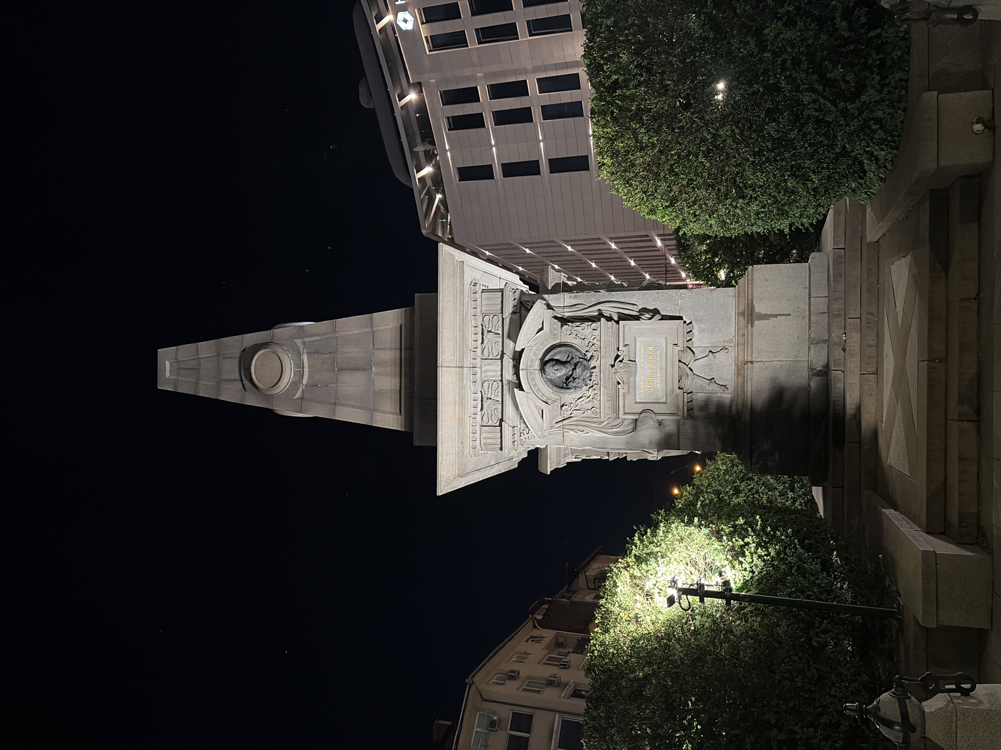 This night-time photograph depicts the Vasil Levski Monument in Sofia, Bulgaria, centrally positioned within an urban environment. The monument comprises a tall, tapering stone obelisk resting upon a substantial, ornate rectangular pedestal. The pedestal's front face features a circular bas-relief portrait of a man, beneath which an inscription plate displays Cyrillic text, clearly including "ВАСИЛ ЛЕВСКИ." The pedestal is adorned with intricate architectural carvings, and multiple stone steps lead to its base. Dense, manicured green hedges frame the monument on both sides, with a bright street lamp illuminating the left-side hedge. In the background, two multi-story buildings are visible: one to the left with a light facade and air conditioning units, and another to the right, a modern light-colored structure featuring dark grid-patterned windows and a rooftop terrace, with a faint "H" logo visible on its upper facade, suggesting a hotel. The sky is dark, characteristic of a night scene, with faint specks of light. No individuals or active interactions are present.