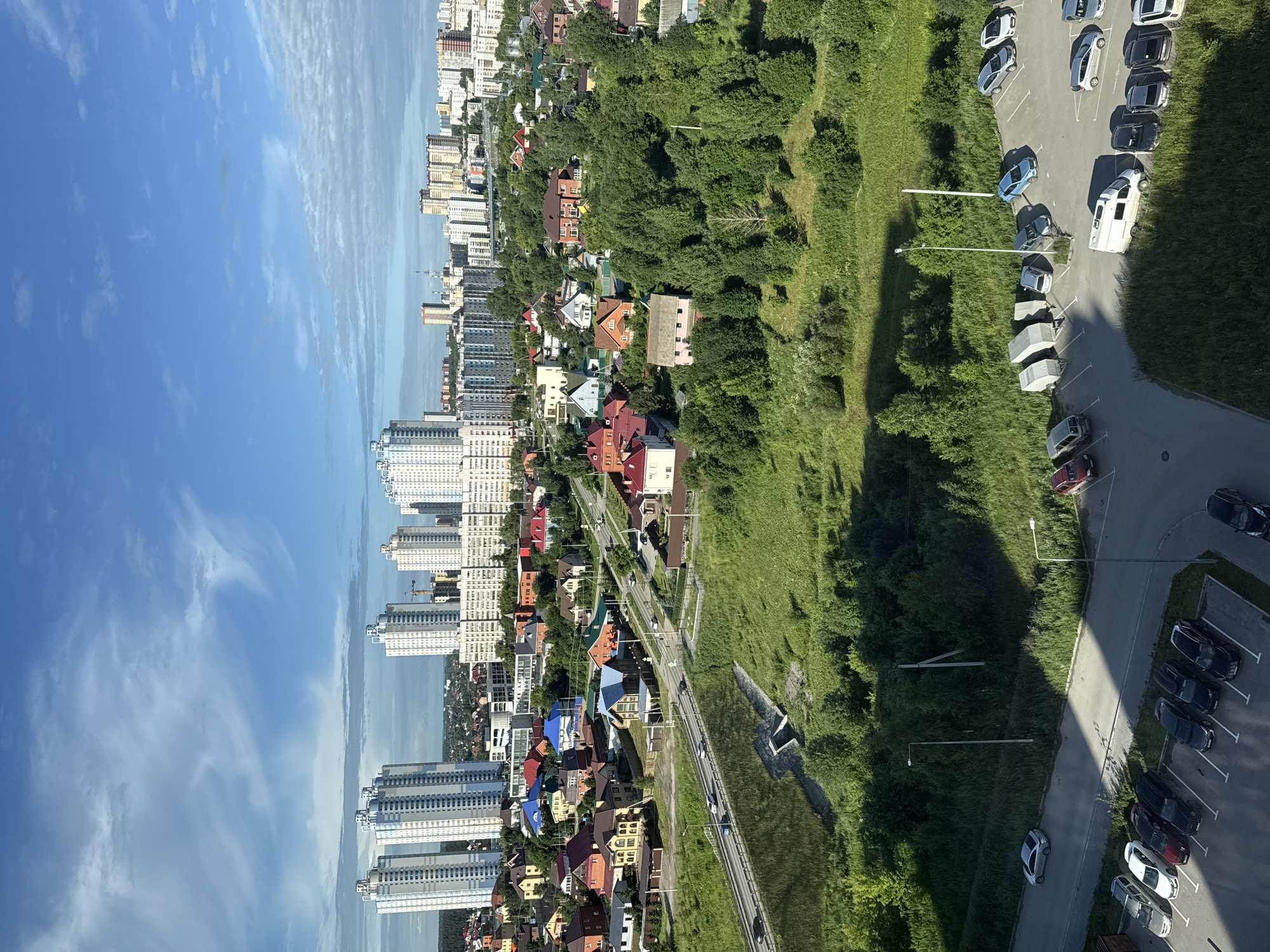 The image depicts an aerial view of a suburban landscape in Perm, Russia. In the foreground, a parking lot contains several vehicles, including cars, vans, and trailers. Beyond the parking lot, a grassy hill slopes down towards a road where cars are driving. Further in the distance, a neighborhood of houses with colorful roofs is visible, surrounded by trees. In the background, tall apartment buildings dominate the skyline, characteristic of urban development. The sky is blue with scattered white clouds, suggesting fair weather. The scene appears to be a blend of residential and urban areas, with a clear division between the lower-density housing and the high-rise buildings.