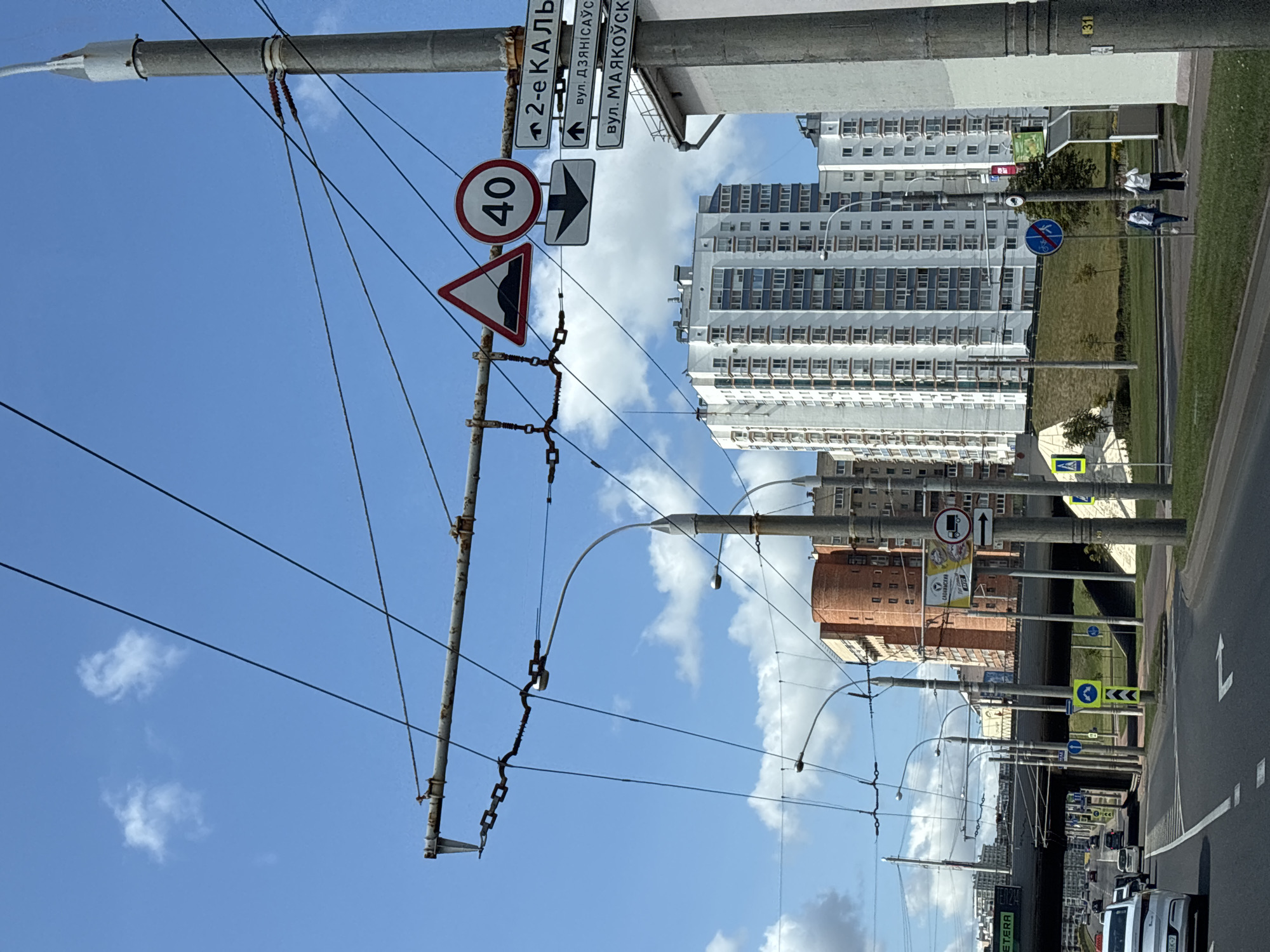 The image shows a street scene in Minsk, Belarus, on a bright, partly cloudy day. In the foreground, a paved road curves to the right. Overhead, a complex network of trolleybus wires crisscrosses the sky, supported by metal poles. Several traffic signs are visible. On a pole on the right, there are signs indicating "2-e KAL'", "вул. ДЗЯНІСАУСКАГА", and "вул. МАЯКОУСКАГА". Also on this pole, a speed limit sign shows "40" and a triangular sign depicts a hump in the road. Further down the street, a large, modern multi-story building with many windows dominates the skyline, with other residential buildings visible behind it. In the distance, an elevated road structure can be seen, with cars driving underneath. Two women are walking on a sidewalk on the right side of the road. The overall impression is a typical urban environment with active infrastructure and modern architecture.
