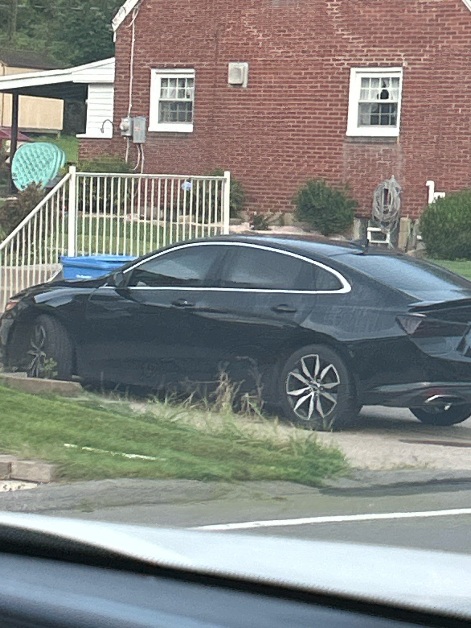 A black sedan is parked on a grassy embankment in front of a brick house. The car has a spoiler on the trunk and multi-spoke wheels. Behind the car, a white fence encloses a yard with a blue recycling bin. The house has white trim around the windows and a white porch roof. A green, patterned object is visible on the porch. The scene appears to be in a residential area.