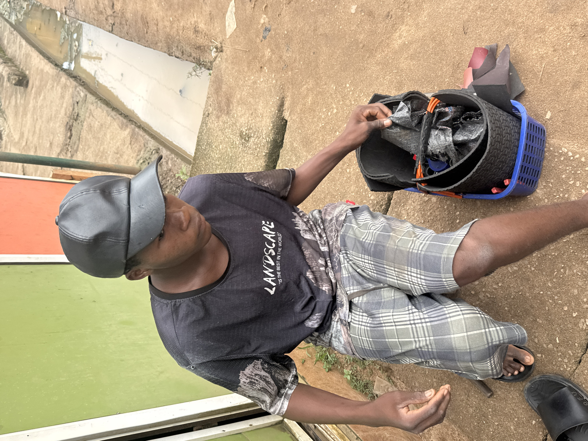 In Ihiagwa, Nigeria, a young person wearing a black cap, a dark t-shirt with the word "LANDSCAPE" printed on it, and plaid shorts sits on a concrete surface. The person is looking down into a blue plastic basket filled with black materials, some of which appear to be rolled-up fabric or rubber. The background features a green wall on the left and a dirty ditch filled with murky water on the right, indicative of an outdoor urban environment. Scattered debris is visible on the ground.