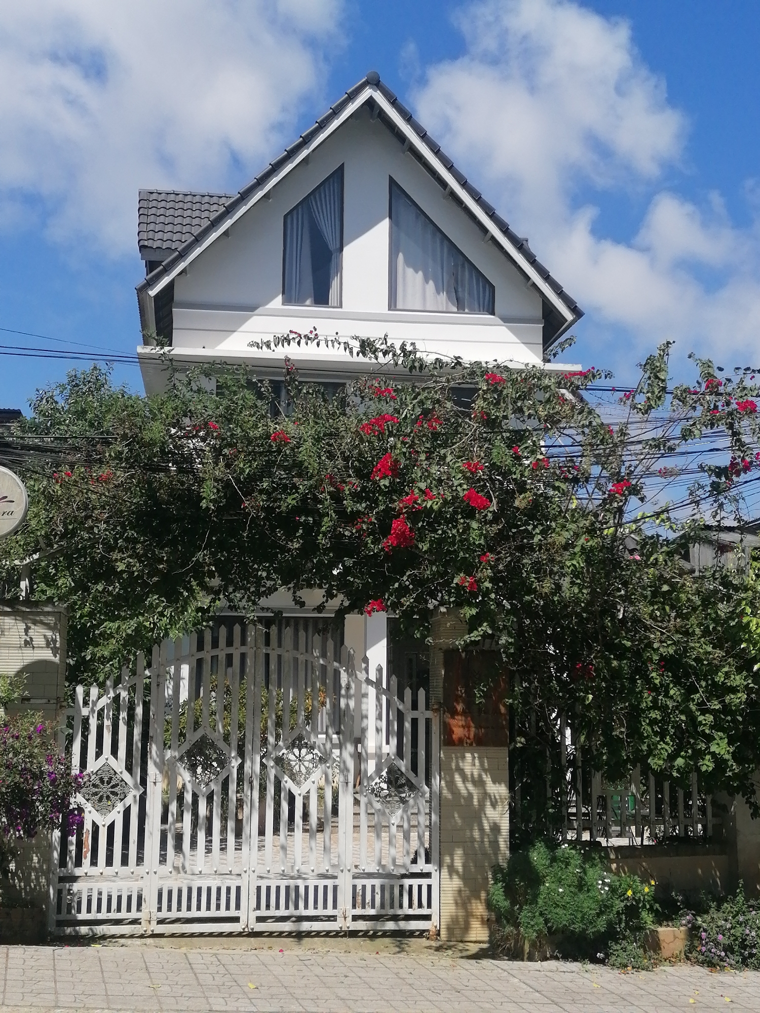 The image depicts a white, two-story house with a grey tiled roof, seen from the front. The house has a distinct gabled section with two triangular windows on the upper floor. A dense vine with red flowers covers a significant portion of the house's facade, extending over a white metal gate. The gate is ornate, featuring vertical bars and diamond-shaped decorative elements. The pathway in front of the gate is paved with grey square tiles. The scene is set against a bright blue sky with scattered white clouds. The location appears to be a residential area in Da Lat, Vietnam, characterized by lush greenery and traditional architectural elements. The image is captured during the daytime.