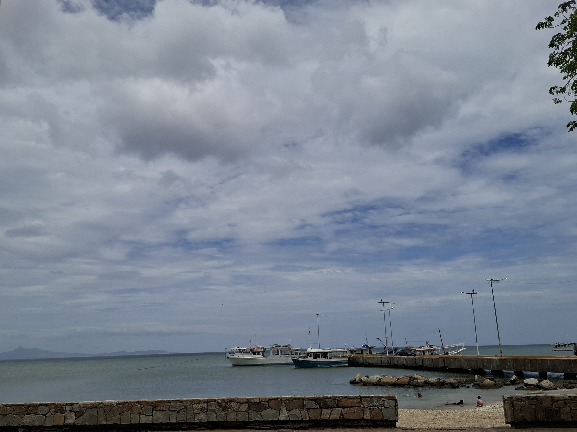 The image shows a coastal scene in Juan Griego, Venezuela. Several boats are docked along a concrete pier, which is lined with street lights. In the foreground, a stone wall borders a small sandy beach where several people are present, some appearing to be playing in the water. The sea extends to the horizon, with a landmass visible in the distance. The sky is overcast with dense clouds.