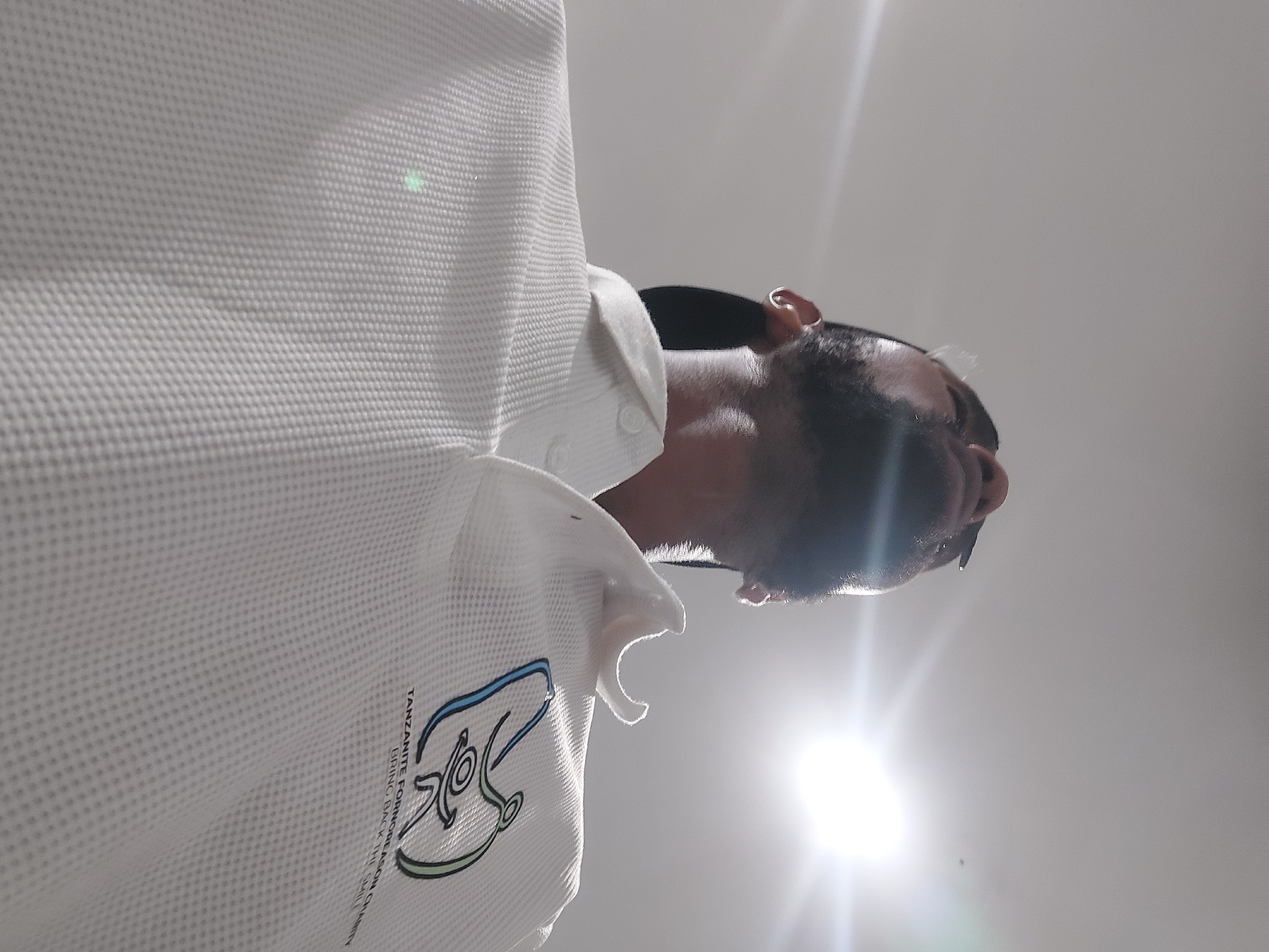 A low-angle shot depicts a man wearing a white shirt with a logo that includes the words "Tanzanite For More Reason Charity Bring Back The Smile", suggesting an association with a Tanzanite-related charity initiative. He has dark hair and a beard. The image is backlit, with a bright light source in the background, making it difficult to discern details of the surrounding environment, but based on the logo and context, it is likely located in Dar es-Salaam, Tanzania. The man appears to be looking down.