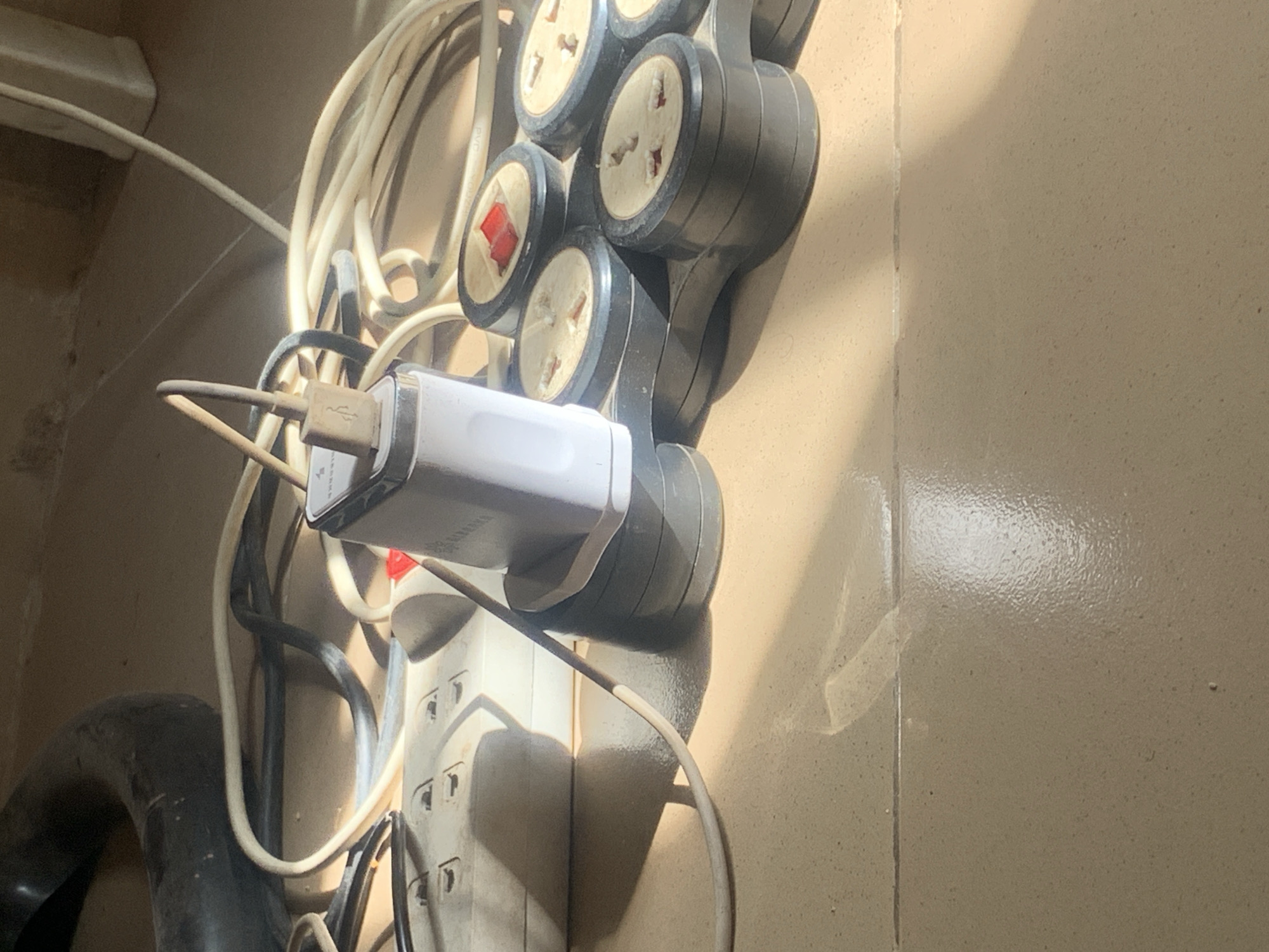 The image depicts a close-up, overhead view of a power strip with multiple outlets and several electrical cords plugged into it. A white USB charger adapter is also plugged into one of the outlets. The power strip and cords are resting on a tiled floor, with sunlight casting strong shadows. The scene appears to be indoors, likely in a residential or office setting. No people are visible in the image. The environment suggests an area with access to electricity. There is no information to place this scene specifically in Jauro Yinu, Nigeria.