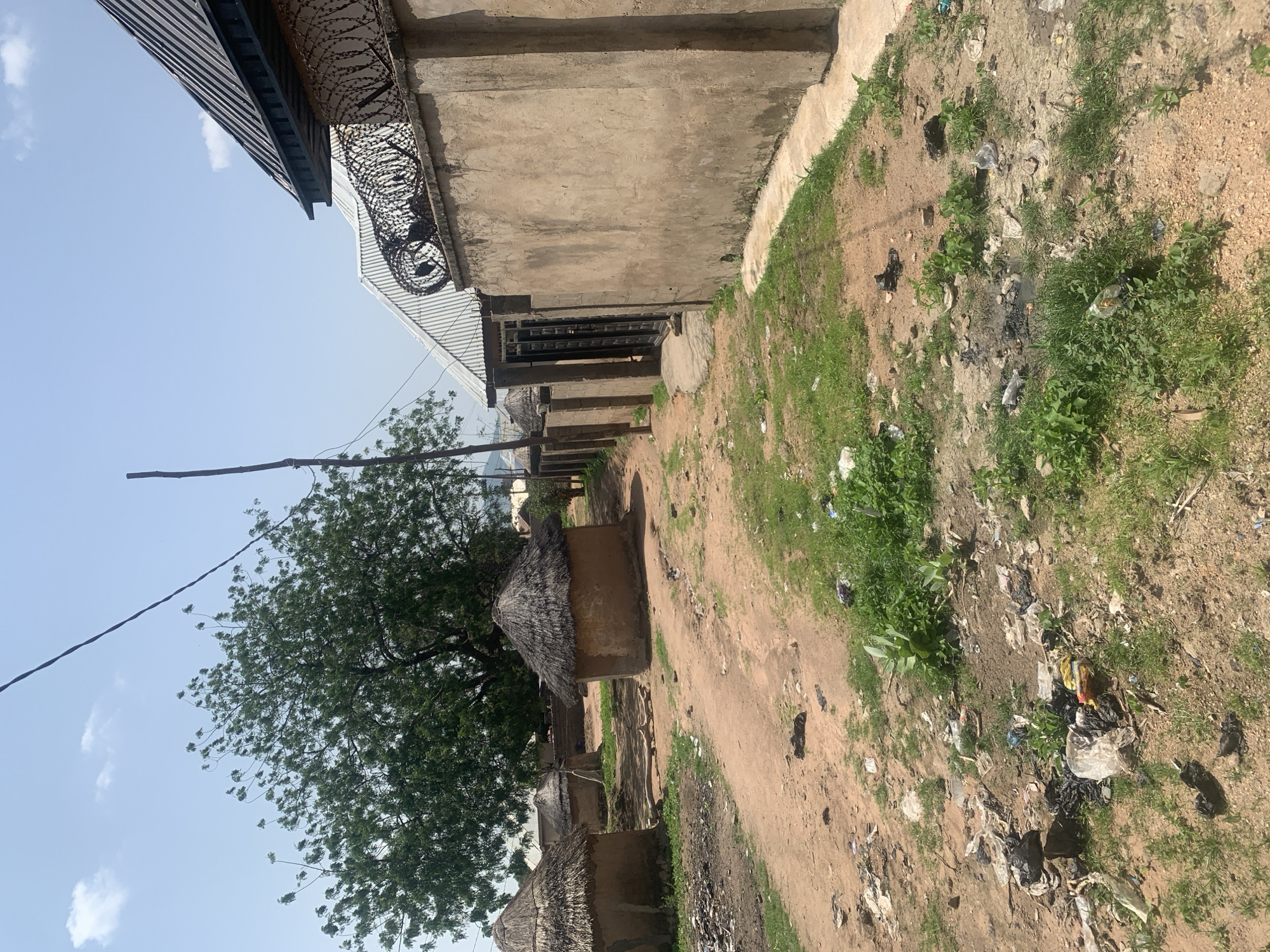 The image depicts a scene in Jalingo, Nigeria, featuring traditional thatched-roof huts with mud walls situated on a dirt path. The path is uneven and scattered with debris, including plastic bags and other refuse. To the right of the path, a concrete wall topped with barbed wire encloses a modern structure with a corrugated metal roof. A black metal gate is visible within this enclosure. In the background, a large, leafy tree stands prominently, with a clear sky overhead. A utility pole and electrical wires are also present in the scene. The overall environment suggests a mix of traditional and modern elements within a somewhat neglected urban setting.