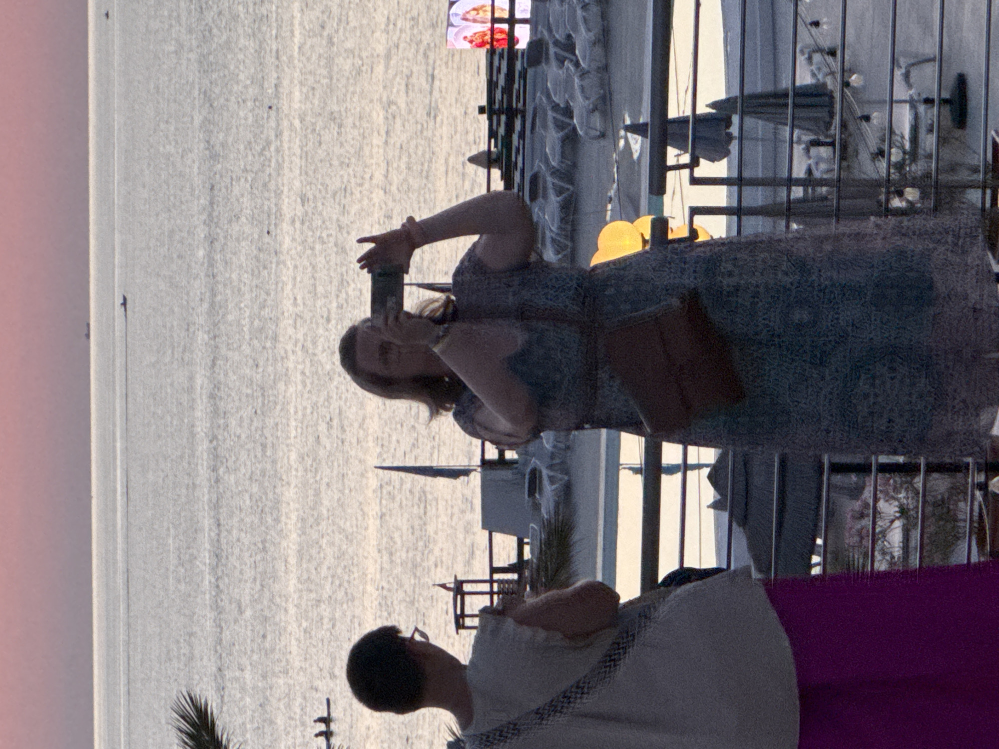 This image depicts a waterfront scene in Isola delle Femmine, Italy, at dusk. Two individuals are visible in the foreground. A person on the left, seen from the back, is wearing a light-colored top and a vibrant purple skirt, with a patterned strap across their shoulder. To the right, a woman is holding a smartphone up to take a photograph. She is wearing a patterned blue dress and has a brown bag slung across her chest.
The background reveals a calm body of water, likely the Mediterranean Sea, with a pinkish-orange sky indicating sunset. Distant boats are visible on the water. Along the shoreline, there appears to be a beach area with lounge chairs and umbrellas, suggesting a resort or public beach. A structure resembling a lifeguard tower or observation post is also present on the beach. Further in the distance, a large illuminated screen displays an image. The overall atmosphere suggests a relaxed evening by the sea.