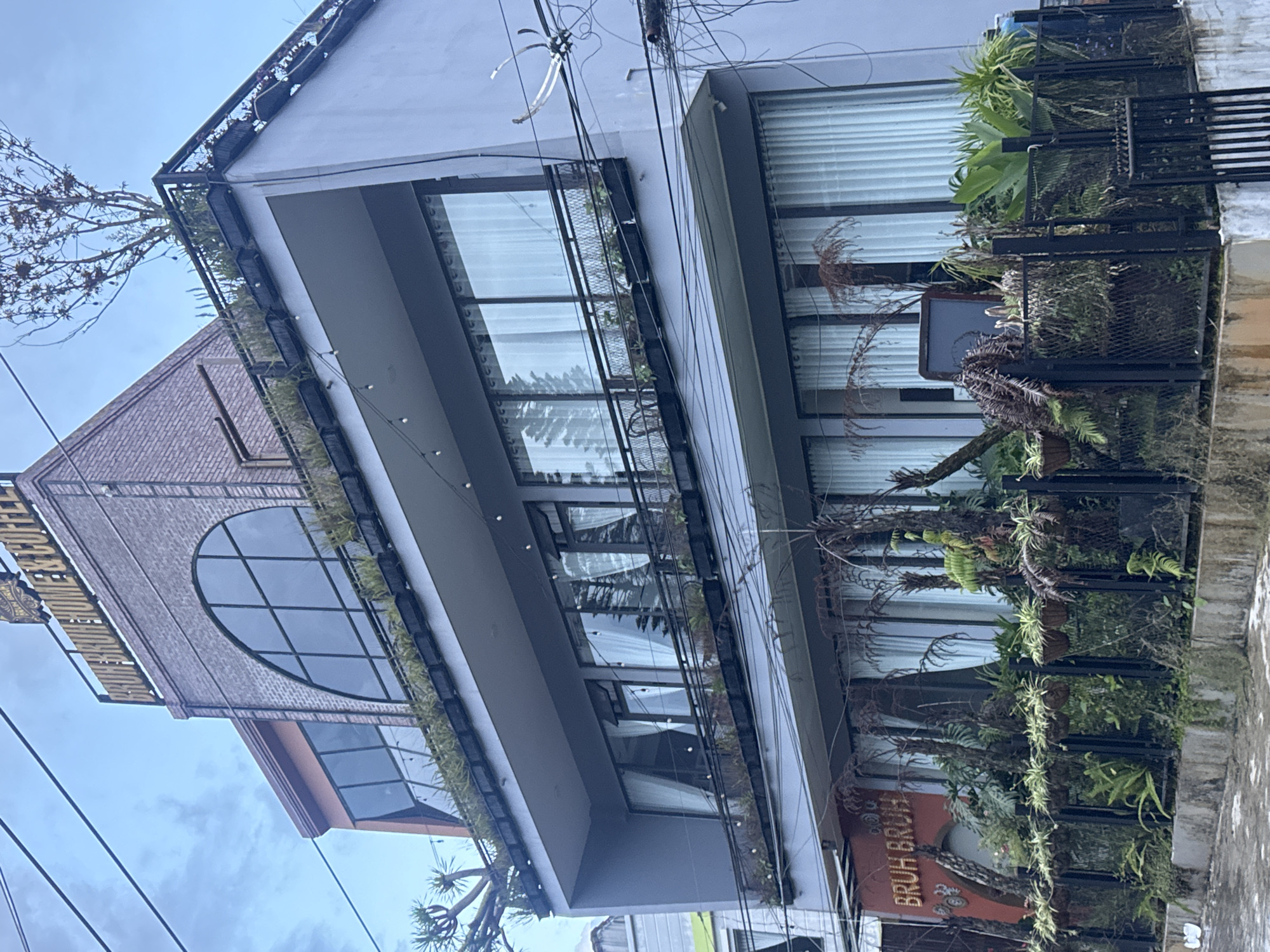 The image shows a multi-story building in Lâm Viên - Đà Lạt, Vietnam. The building has a modern architectural design with large windows and balconies adorned with plants. On the top floor, a brick facade features a large arched window, and the building's name, "BRUH BRUH HOME & COFFEE," is visible. The ground floor has a similar facade with a red-orange sign that says "BRUH BRUH." The building is surrounded by lush greenery and plants, suggesting a garden or cafe setting. Overhead, power lines crisscross the frame, indicating an urban environment. The sky is overcast, and the overall scene depicts a commercial establishment in a city known for its natural beauty.