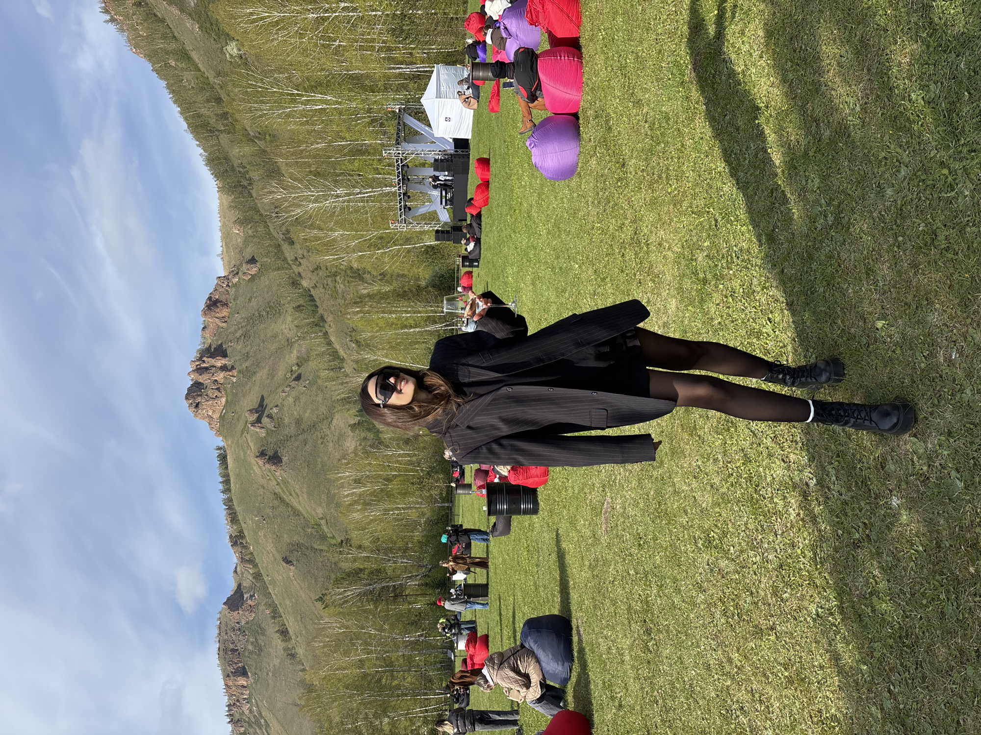 The image shows a woman posing in an outdoor setting. She wears a black pinstripe blazer, black tights, and black boots. She also wears sunglasses. She stands on a grassy field where an outdoor event seems to be taking place, with a stage set up in the background and people seated on colorful beanbags and black barrels. The landscape includes a mountain ridge and trees.
While the location is not explicitly stated as Krasnoyarsk, Russia, the presence of such a mountainous landscape near an outdoor event suggests a location within or near Krasnoyarsk, which is known for its surrounding natural landscape.