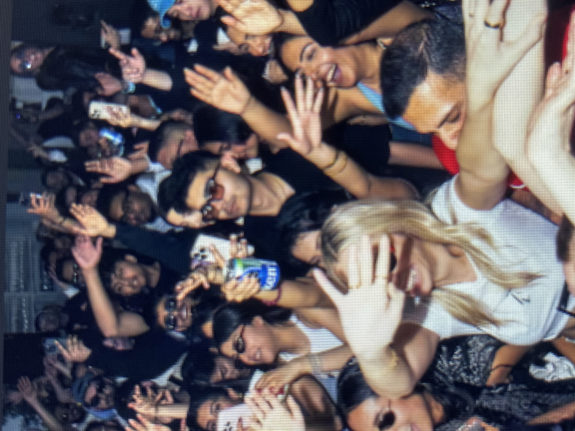 The image depicts a crowded indoor event, possibly a concert or party, in an urban setting. A diverse group of young adults are visible, many with their hands raised in the air, seemingly enjoying themselves. Some individuals are holding up smartphones, likely recording or taking pictures. One person in the center is holding a blue and green can, possibly a beverage. The lighting is dim, contributing to a lively atmosphere. The faces of the attendees show expressions of excitement and engagement. The background is indistinct, suggesting a busy and energetic environment.