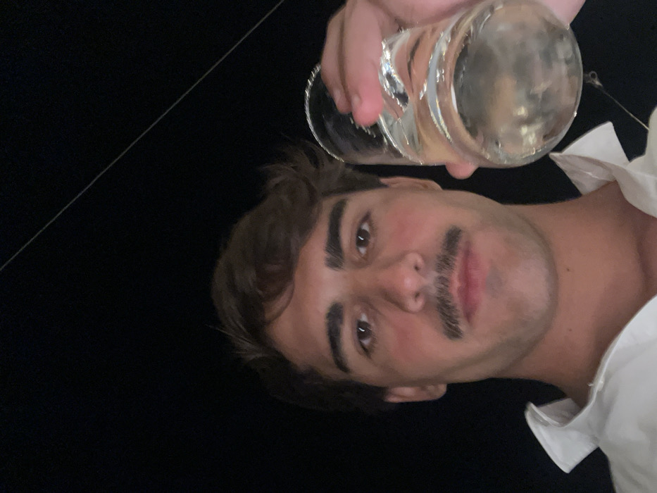 A dark, upward-angled selfie shows a man with brown hair and a mustache holding a glass of liquid above his head. He wears a white collared shirt, which is visible at the right side of the frame. The background is solid black, save for a faint diagonal line in the upper left quadrant.