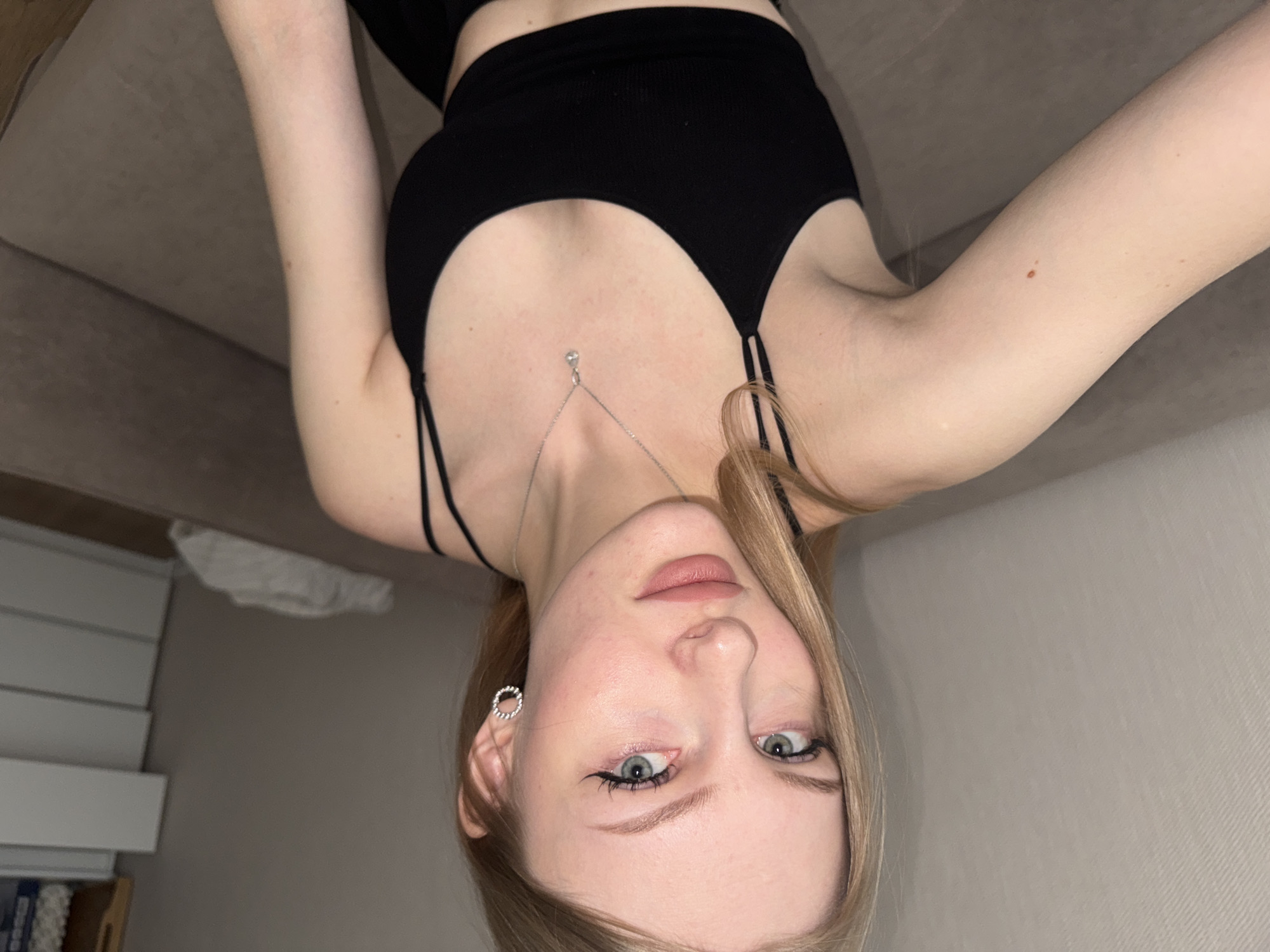 A young woman with fair skin, light blonde hair, and blue eyes is the primary subject of the image. She is wearing a black tank top with thin straps and a silver necklace with a pendant. Her left ear is visible, adorned with a small, round earring. She appears to be in an interior setting with a beige couch visible in the background. The image seems to be taken from an upside-down perspective, as the background elements are oriented in reverse. Elements in the background include white cabinets, and some books. The image does not include information to determine the location of Perm, Russia.