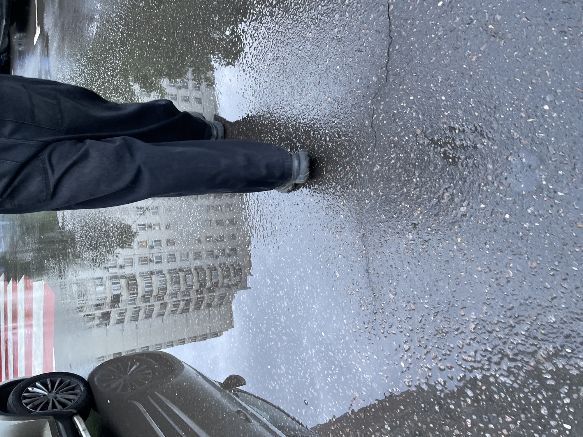 The scene depicts a street in Moscow, Russia on a rainy day. A person wearing dark pants and shoes stands on a wet asphalt surface. Reflections of a building and red and white striped poles are visible in the puddles. The rear tire and part of a dark-colored car are also present in the frame. The ground is covered in water droplets.