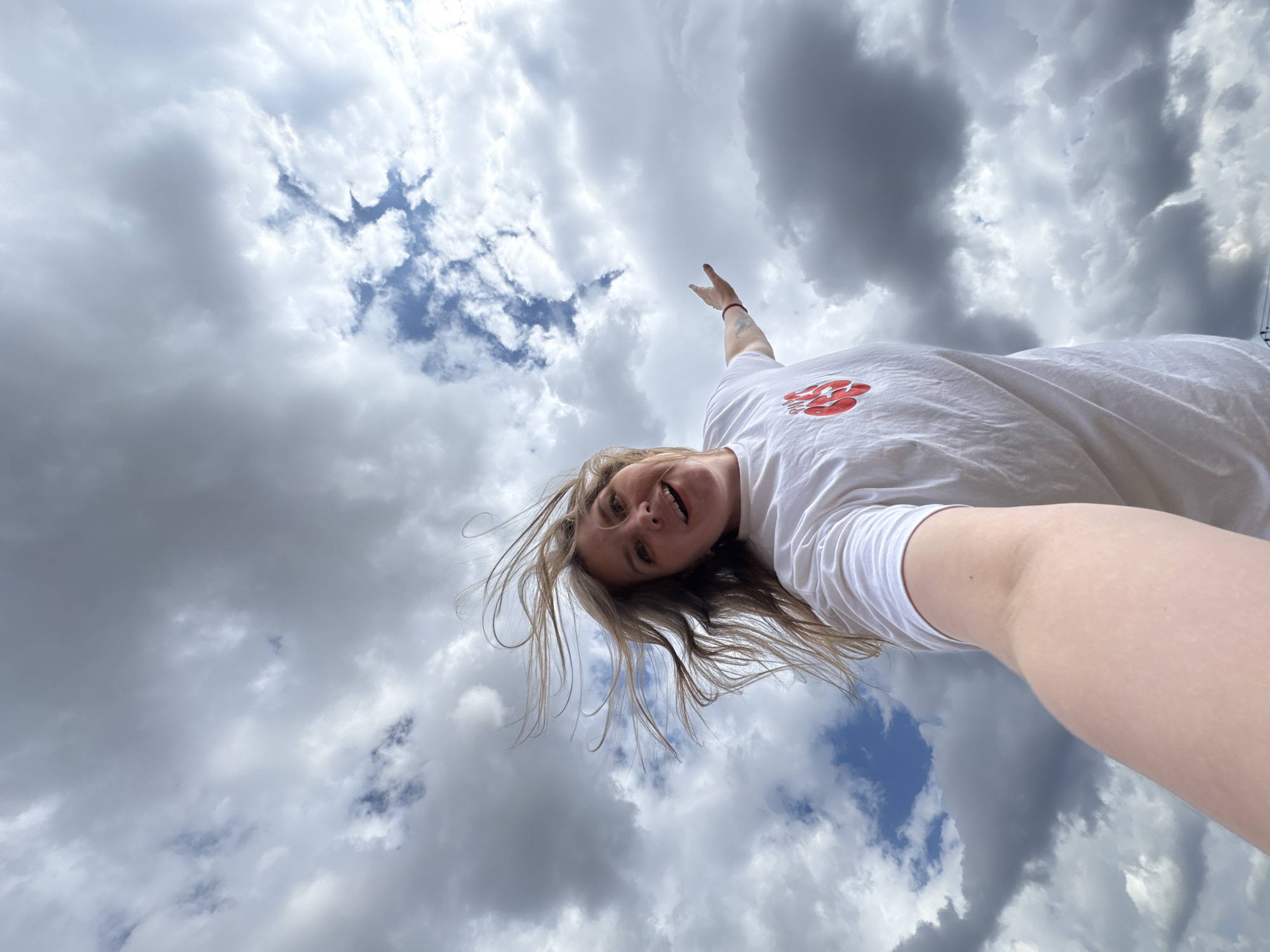 The image, captured in Gornyi (Dvurechenskoe S/P), Russia, presents a unique low-angle selfie of a smiling woman set against a dynamic cloudy sky. The subject is a young Caucasian woman with light brown hair that appears swept upwards and outwards, suggesting movement or wind. She gazes downwards into the camera with an open-mouthed smile, her head slightly tilted, conveying a cheerful and playful demeanor. She wears a plain white t-shirt adorned with a red graphic logo on the chest; this logo features stylized letters, possibly "ST," followed by two interconnected circular shapes. Her right arm is raised, with her hand pointing directly upwards into the vast expanse of the sky.
The background is entirely composed of varying cloud formations, ranging from bright white to darker grey, interspersed with patches of clear blue sky. This overhead perspective creates an illusion of the woman floating or looking down from above the clouds. The bright, natural illumination indicates that the scene takes place during the daytime. No specific landscape or architectural features are visible, emphasizing the expansive sky and the woman's playful interaction with it.