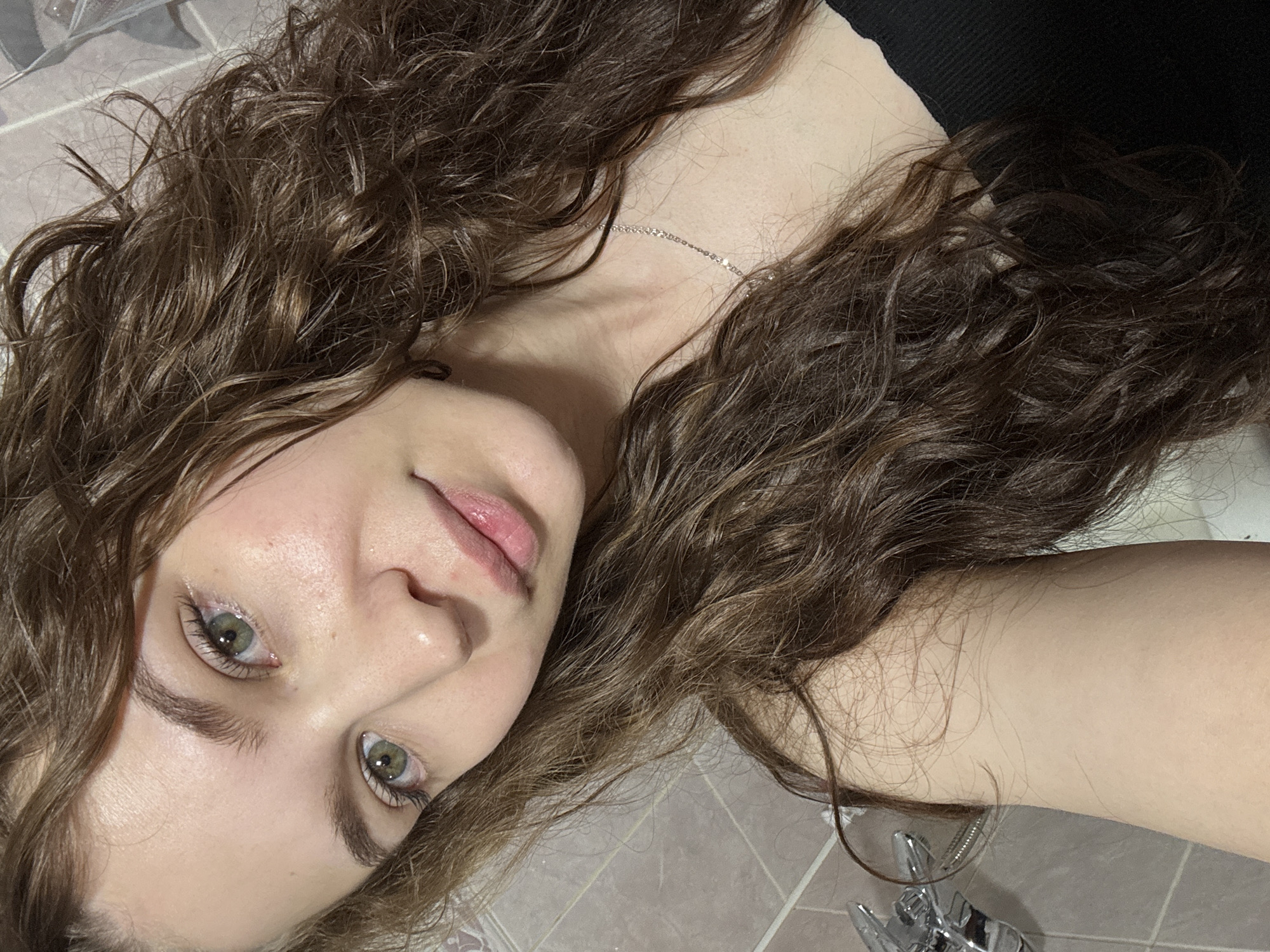 The image presents a close-up portrait of a young individual, likely female, with fair skin, striking green eyes, and full, lightly glossed lips. Her expression is neutral to slightly serene, and her gaze is directed straight at the camera. She has long, voluminous, curly brown hair that cascades around her face and shoulders, appearing textured and slightly damp. A delicate silver chain necklace is visible around her neck, resting on her collarbone. A black garment, possibly a top or strap, can be seen near her right shoulder.
The person appears to be lying down or reclining, captured from a slightly overhead perspective. The background consists of a light-colored, possibly beige or light grey, tiled surface, suggesting an indoor setting such as a bathroom or a room with tiled flooring. A small, metallic object, resembling part of a fixture like a faucet, is partially visible in the lower right corner of the frame. The lighting is soft and even, indicating an indoor environment without harsh direct light. No text is visible in the image. This scene takes place in Chaikovsky, Russia.