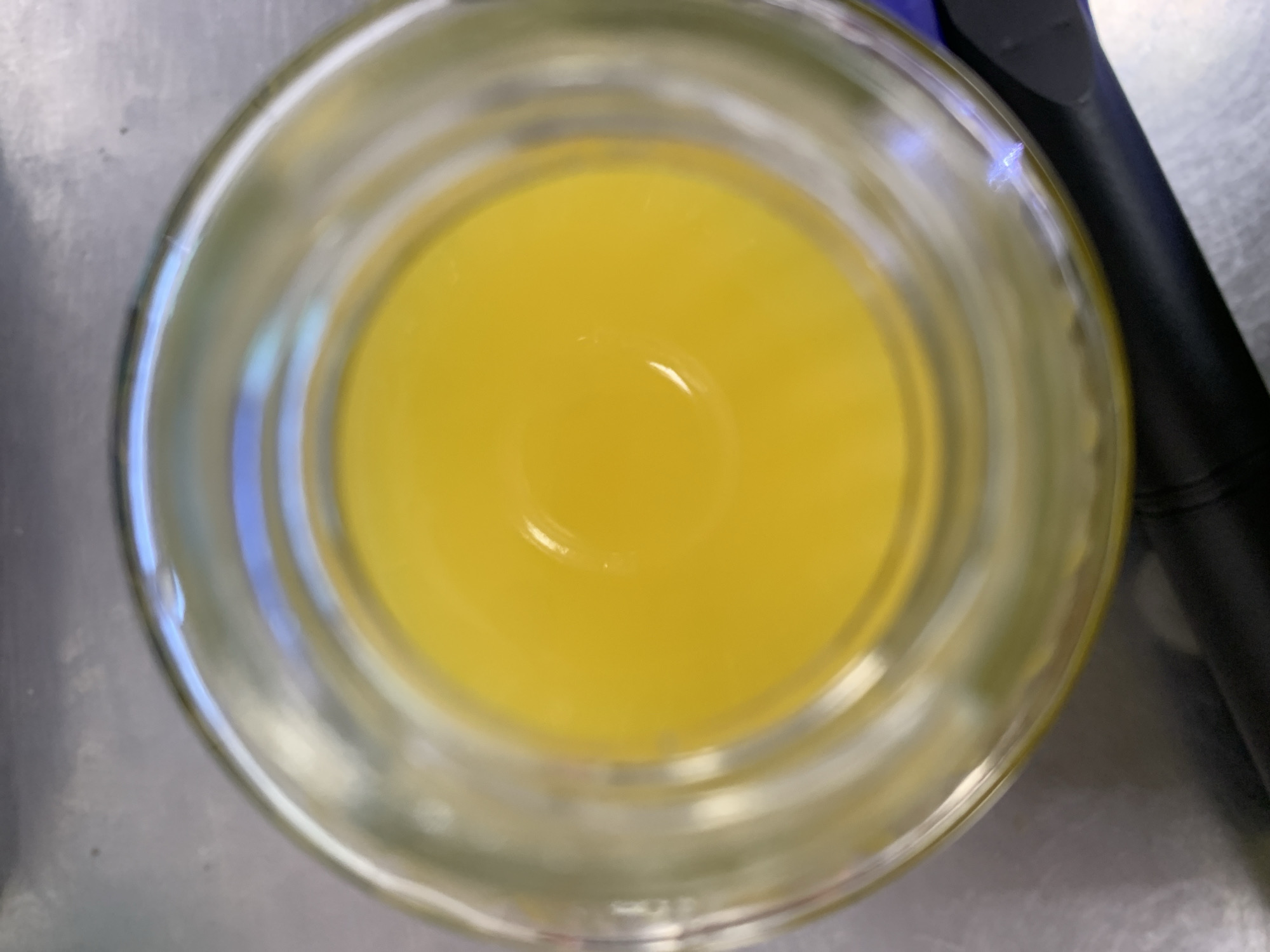 This media file is a top-down, close-up view of a clear glass jar filled with a bright yellow liquid. The liquid appears to be viscous and has a smooth surface with a slight ripple or indentation in the center. The glass jar has a thick rim and is placed on a metallic surface that has a brushed or textured finish. A portion of a dark object, possibly a tool or utensil with a black handle, is visible to the right of the jar. The lighting suggests an indoor setting, and there are no external cues to determine the time of day or weather. No text is visible in the image. The overall impression is a simple, still life composition focusing on the color and texture of the liquid within the container.