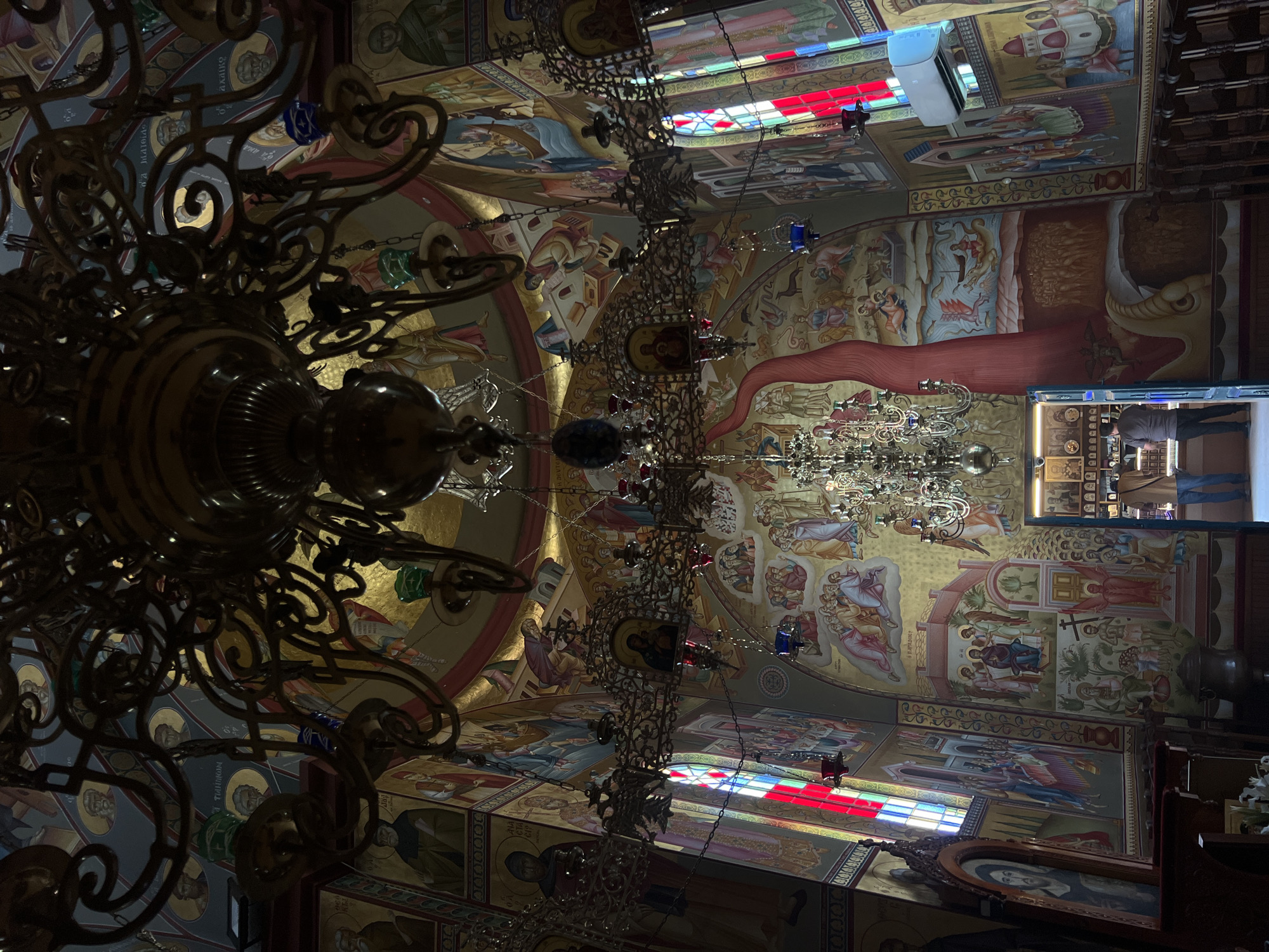 The image depicts the interior of a church, likely an Orthodox Christian church, featuring extensive religious murals adorning the walls and ceiling. A large, ornate chandelier hangs prominently in the foreground, with another smaller chandelier visible further back. Stained-glass windows with red and blue panes are visible on the left, casting colored light into the space. In the background, a doorway leads to an area with icons displayed on shelves. Two individuals, one wearing a brown shirt and the other a gray shirt, are standing in the doorway, appearing to examine the icons. The church appears to be located in Emek HaYarden Regional Council, Israel, given its architectural style and prevalent religious iconography often found in the region.