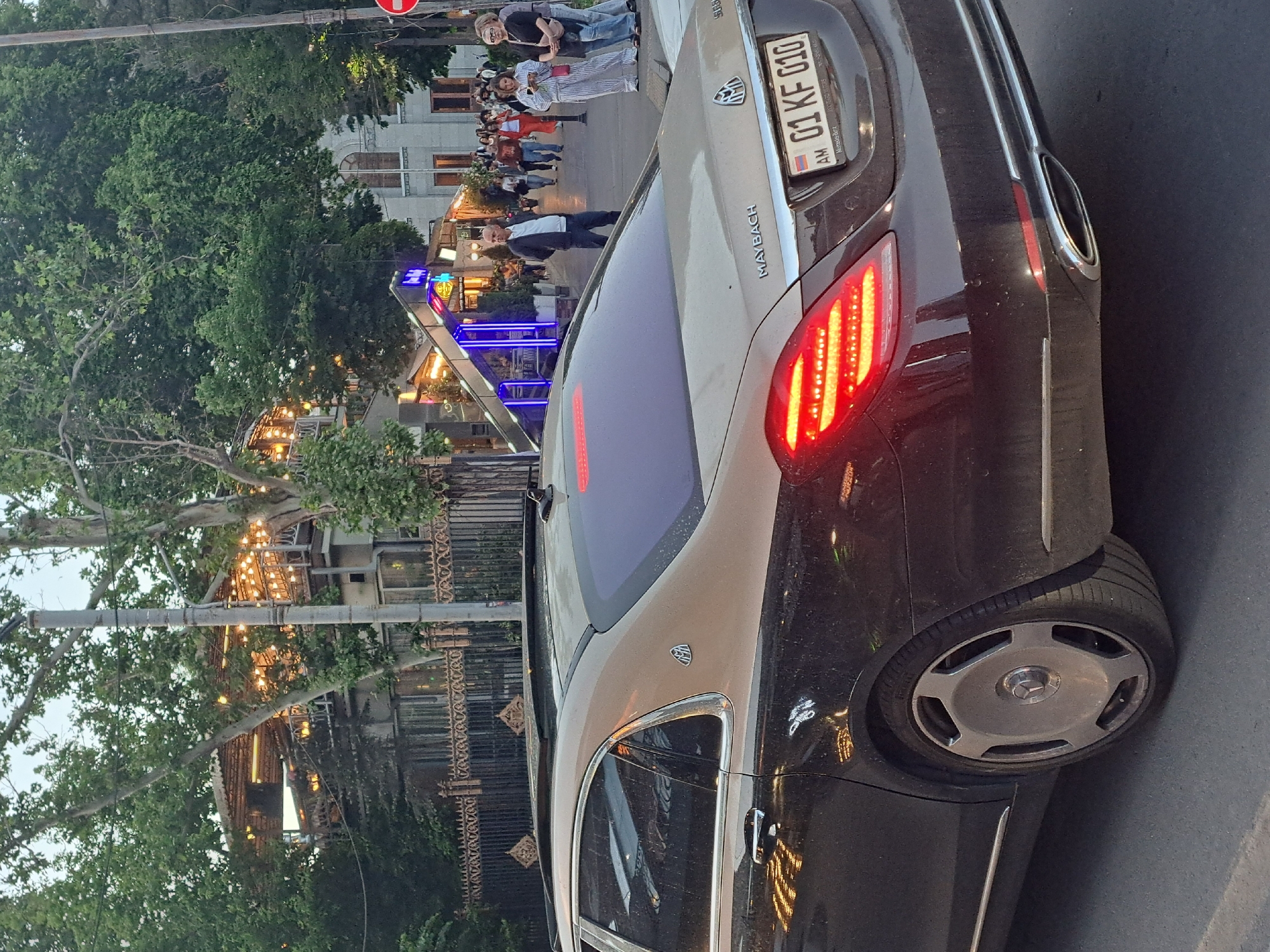 The image shows the rear of a two-tone (tan and black) Maybach car parked on a street in Yerevan, Armenia. The license plate reads "AM 01 KF 010." A group of people are walking along the sidewalk in the background. There are buildings and trees lining the street, and a restaurant or cafe is visible with blue and purple lights. Street lighting adds to the ambiance.