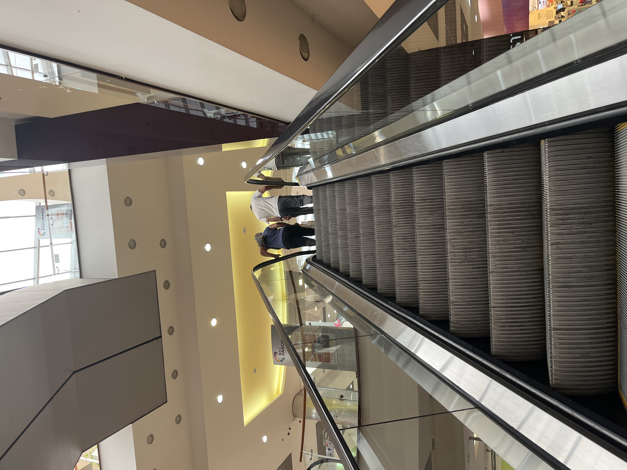 This image, rotated 90 degrees clockwise, captures an indoor scene dominated by a long escalator moving downwards. Two individuals are visible on the escalator, positioned towards the top of the frame. One person, wearing a white shirt and dark trousers, stands upright. Slightly ahead of them, another person, wearing a dark blue top and dark trousers, also stands on the escalator steps.
The setting appears to be a modern, multi-story commercial building, likely a shopping mall, located in Pleven, Bulgaria. The architecture features clean lines, light-colored walls (beige and off-white), and a ceiling with numerous recessed circular lights providing illumination. A prominent dark brown structural beam crosses the upper part of the frame, underneath a reflective glass surface and a white ceiling. Glass railings with metallic handrails line both sides of the escalator, reflecting parts of the interior and creating a sense of spaciousness.
Bright ambient light suggests it is daytime, with large windows or skylights visible on the far left of the original image, contributing to the overall brightness. The escalator steps are a textured grey, and the side panels are silver-toned metal.
Visible text on a sign adjacent to the escalator's glass railing reads "PANORAMA," strongly suggesting the location might be Panorama Mall. The mood of the scene is calm, with the individuals simply riding the escalator as part of their routine transit within the building.