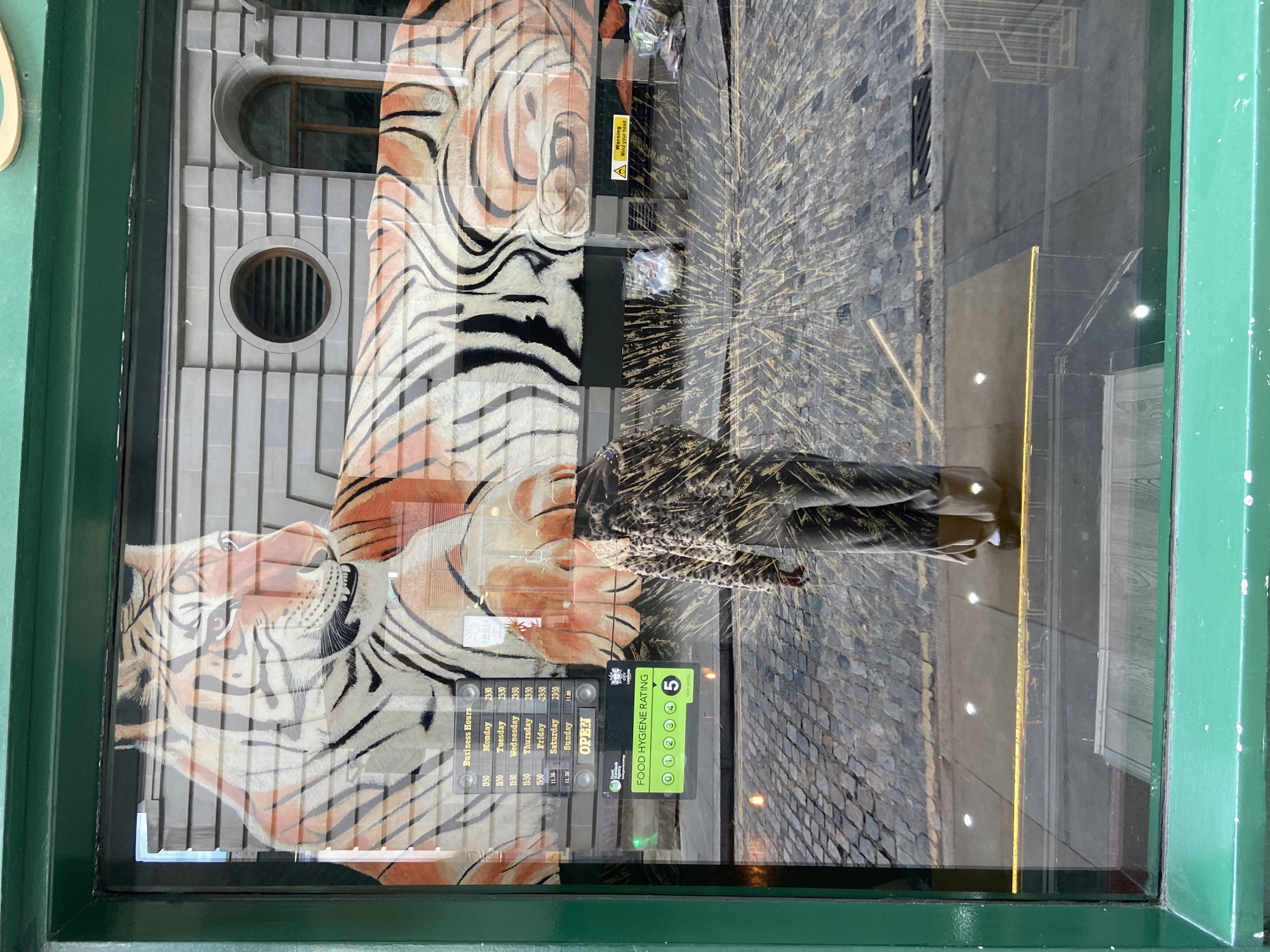 The image captures an urban street scene reflected in a shop window, which itself features a large, stylized graphic of a tiger. The tiger, depicted with distinctive black, white, and orange stripes, dominates the left side of the window, with its head looking towards the left.
The window's reflective surface shows a person walking away from the viewer on a cobblestone pavement, wearing a dark, patterned coat. Further reflections reveal a multi-story building with a light-colored, ornate stone facade and various window styles, including arched and circular designs, characteristic of architecture found in London, United Kingdom.
Several signs are affixed to the inside of the window. A "Business Hours" sign lists opening times from Monday to Sunday, mostly 11:30 to 23:30, with Friday starting at 16:30 and Sunday showing 11:30 to 11:00 (likely a typo for 23:00). An "OPEN" sign is prominently displayed. Below the business hours, a "FOOD HYGIENE RATING" sign, issued by "LONDON," indicates a "5 VERY GOOD" rating. A small yellow triangular sign with black text warns: "Warning Mind your head." The entire scene is framed by a green window casing. The setting appears to be daytime, with ambient light illuminating the reflections and signs.