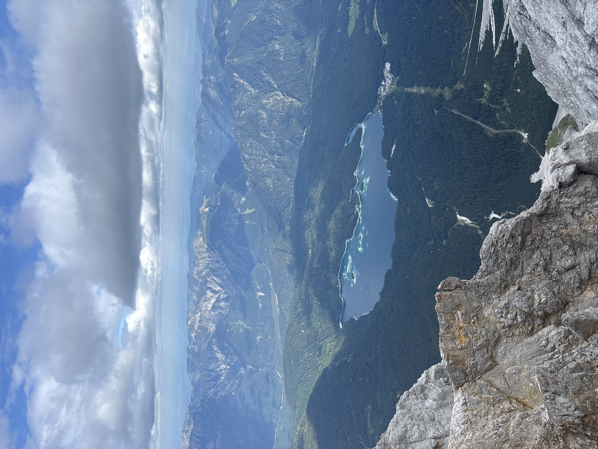 This is a high-angle, wide shot of the Eibsee lake and surrounding mountains, likely taken from the Zugspitze, near Garmisch-Partenkirchen, Germany. The sky is partly cloudy, with large, dramatic gray clouds contrasting with patches of blue sky. In the foreground, a rugged, rocky mountain face dominates the lower left portion of the frame. Below this, the expansive Eibsee lake, a vibrant turquoise blue, is visible. The lake is dotted with small, green islands. The shores of the lake are lined with dense, dark green coniferous forests. In the distance, layers of majestic, blue-hued mountain ranges recede towards the horizon. A ski lift or cable car line is visible ascending a mountainside towards a small cluster of buildings near the lake. The overall scene depicts a vast, natural alpine landscape.