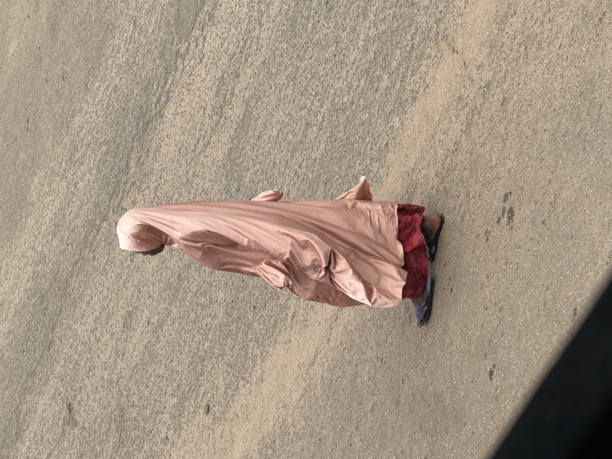 In Bukuru, Nigeria, a person wearing a rose-colored abaya and red skirt walks on a paved road. The abaya is made of a satin-like material and has voluminous sleeves. The person wears dark flat sandals. The road surface is a mixture of asphalt and gravel. The lighting suggests it is daytime.