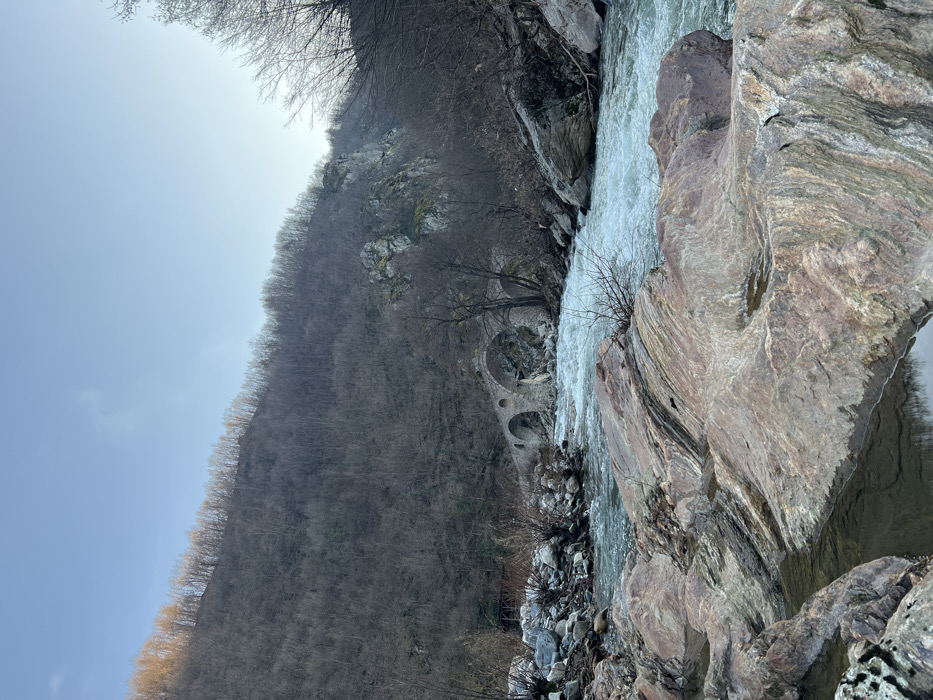 The image depicts a natural landscape in Diadovtsi, Bulgaria, featuring a river valley under a clear, light blue sky with minimal cloud cover. In the foreground, large, multi-colored rock formations with reddish-brown, grey, and dark green hues extend across the bottom, containing shallow pools of still water. A fast-flowing river, exhibiting turbulent, light blue and white rapids, traverses the mid-ground from the lower right to the center, leading towards a historical stone arch bridge.
The bridge, constructed from dark grey and brown stone, spans the river with at least three visible arches, showing signs of age and weathering. It connects the riverbanks. The surrounding environment consists of steep hillsides covered predominantly by deciduous trees and shrubs, currently bare of leaves, suggesting a winter or early spring season. The foliage on the upper ridge of the hill on the far left appears slightly illuminated with a yellowish-orange hue, contrasting with the generally muted tones of the bare trees. Exposed rocky outcrops are visible on the slopes, particularly on the right side of the valley. No individuals or anthropogenic activity are present within the scene.