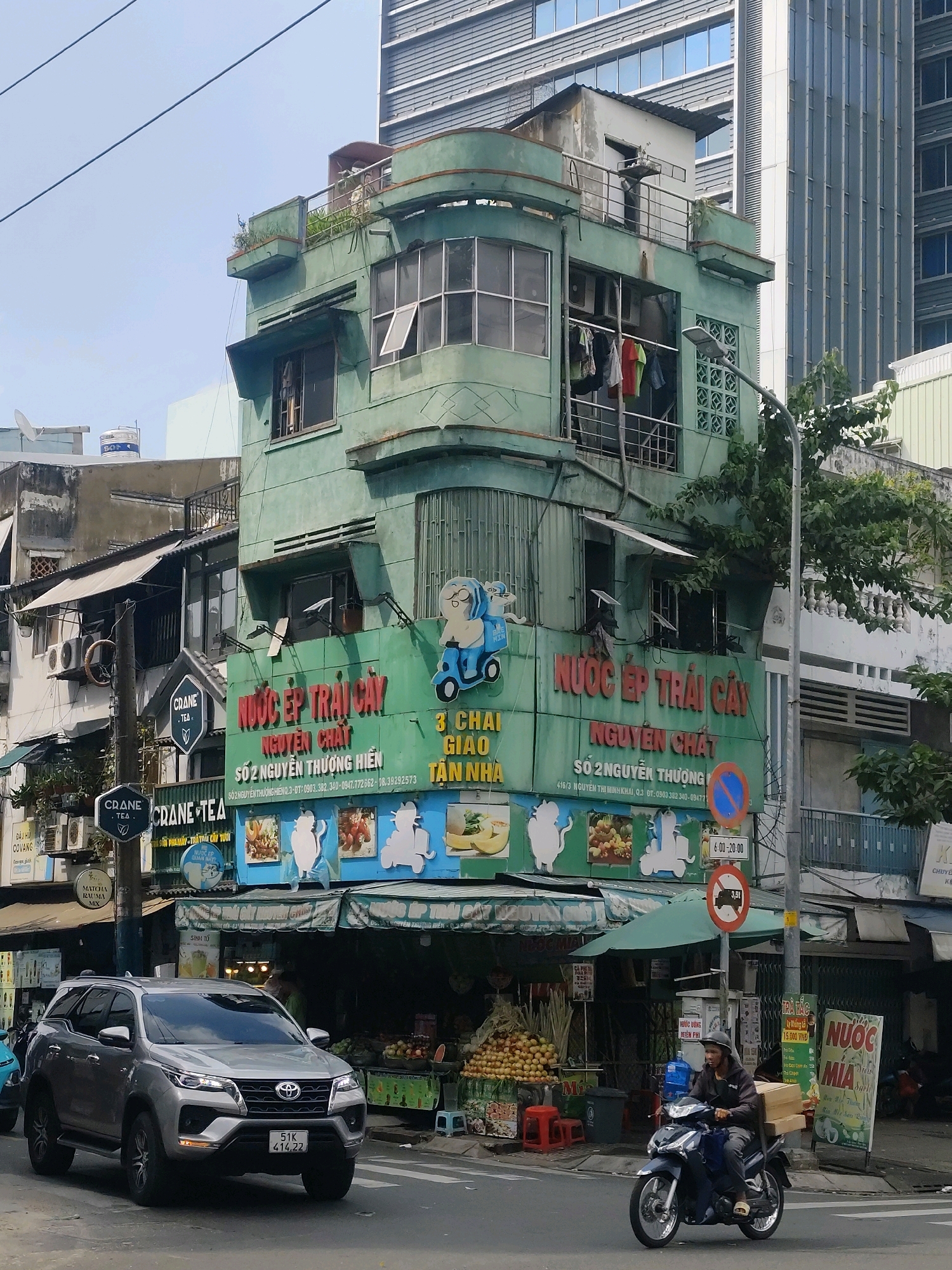 The image shows a street scene in Ho Chi Minh City, Vietnam, on a sunny day. In the foreground, a silver Toyota Fortuner SUV is driving on the road. To the right, a person is riding a dark-colored motorcycle, carrying a load of wooden planks. The background features a weathered, pale green, multi-story building with balconies and windows, adorned with signage in Vietnamese, including "NƯỚC ÉP TRÁI CÂY NGUYÊN CHẤT" (Pure Fruit Juice) and "TRÀ CÁC LOẠI" (All Kinds of Tea). Adjacent to this building is a modern, tall, grey-glass skyscraper. A street light and traffic signs are visible on the right side of the road. The street is lined with small shops and market stalls selling fruits and beverages. The sky is clear and blue.