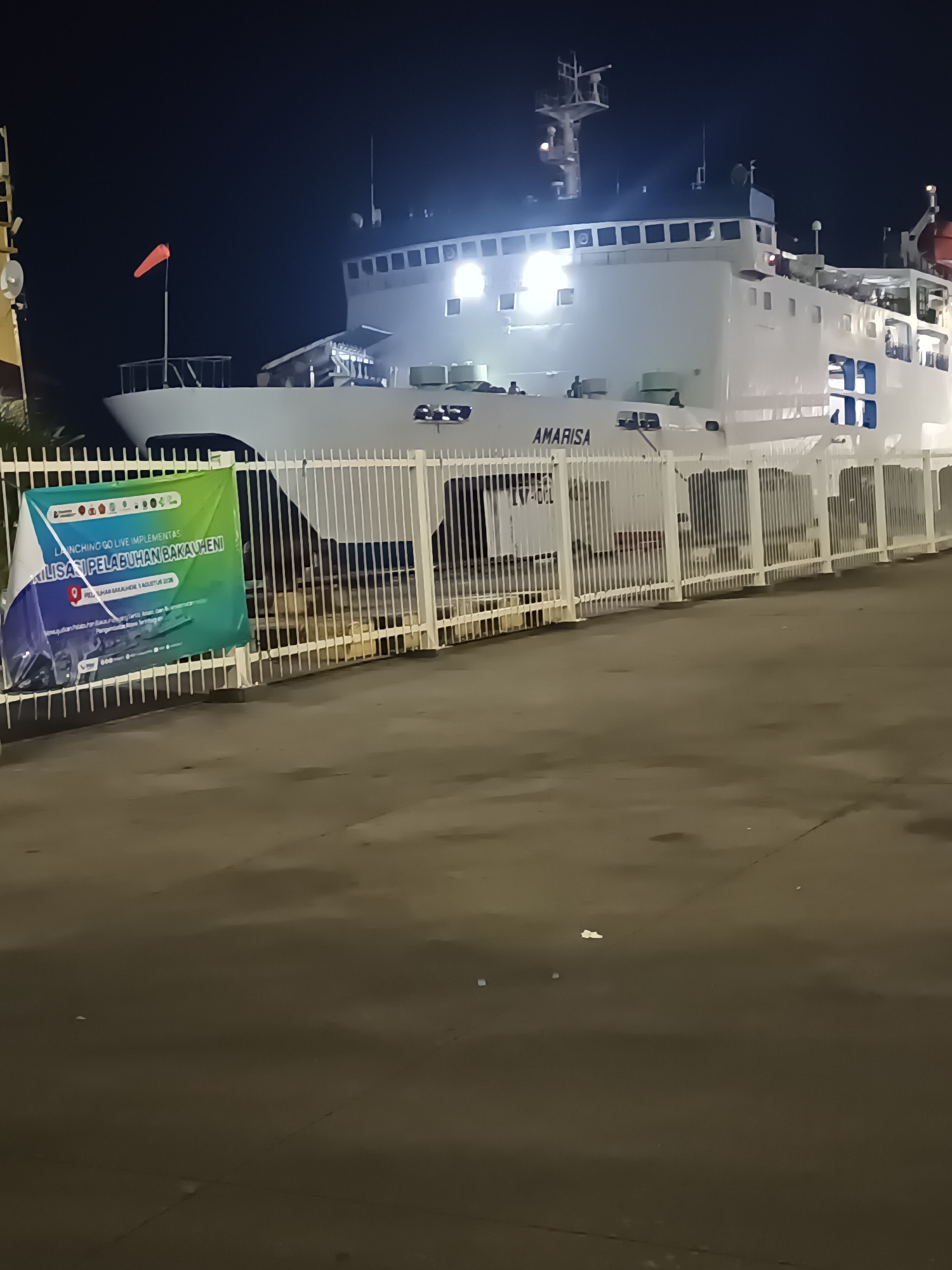 The image shows a large white ferry named "AMARISA" docked at the Port of Bakauheni, Indonesia, at night. The ferry is brightly lit, illuminating its white hull and the upper decks. A green and blue banner is displayed on a white fence in the foreground, partially obscuring the lower portion of the ferry. The banner contains text in Indonesian, including "LAUNCHING GO LIVE IMPLEMENTAS" and "PELABUHAN BAKAUHENI". The ground in front of the fence appears to be asphalt. The overall scene depicts a port facility at night, with the ferry being the primary subject.