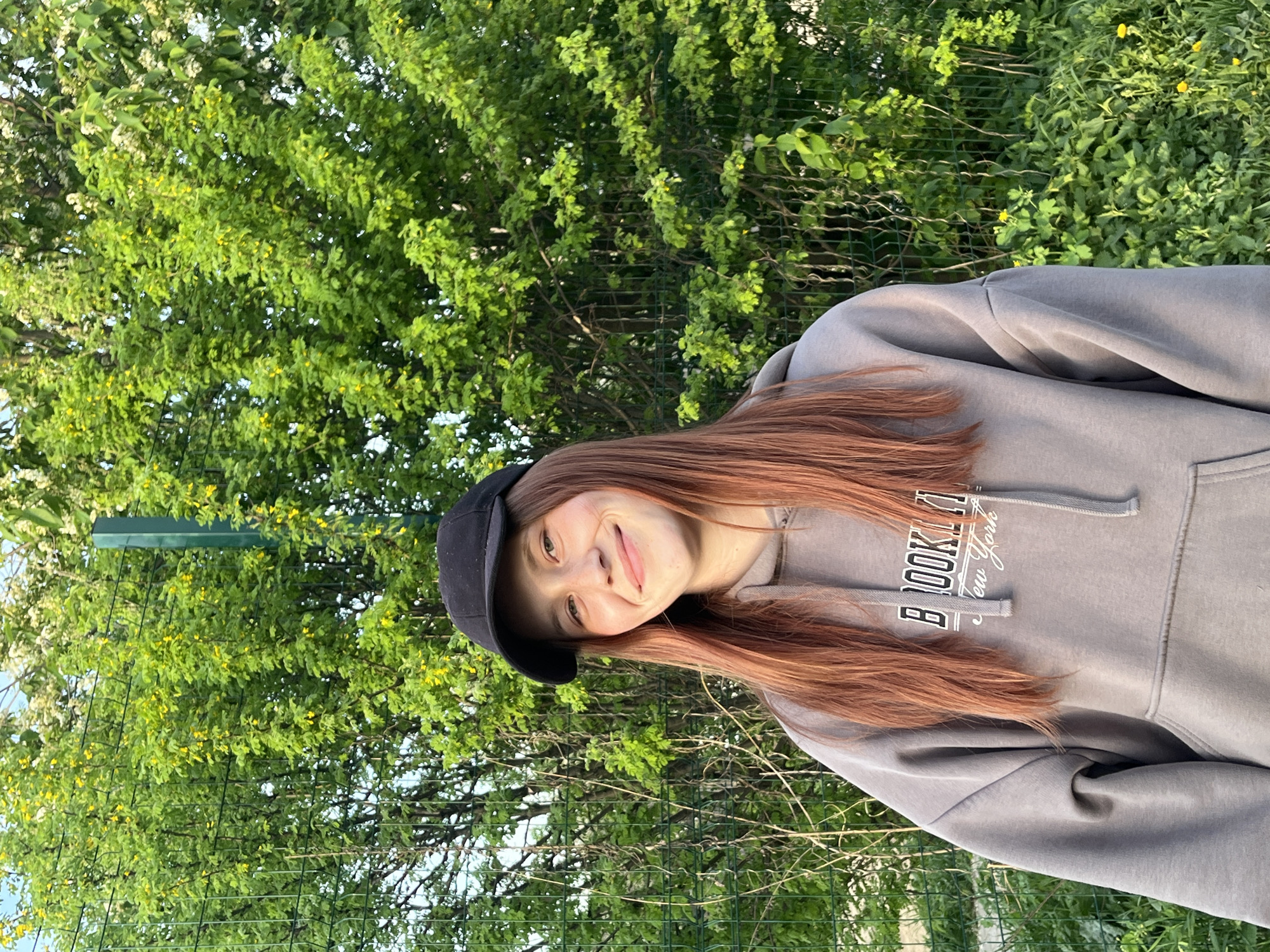 A young woman stands outdoors, wearing a gray hooded sweatshirt and a black cap. Her reddish-brown hair falls around her shoulders. She is smiling slightly and looking at the camera. Behind her, there's a dense green background of trees and vegetation, partially obscured by a chain-link fence. Yellow flowers are scattered among the green foliage. The location details (city of Фоки, Russia) were not discernible from the provided image and hints.