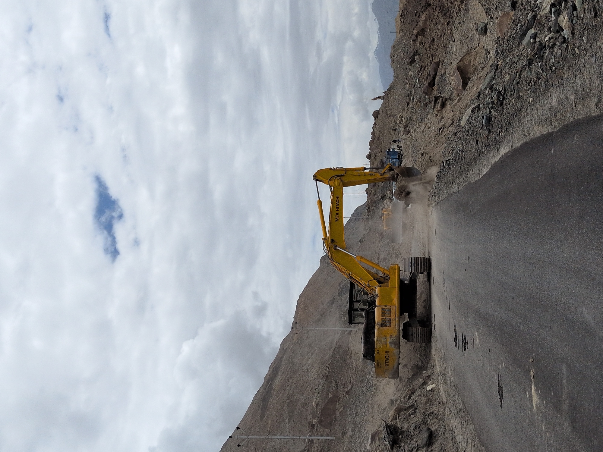 A wide-angle, eye-level shot captures a construction scene on a road in Panamik, India, under a cloudy sky. A large yellow excavator, identified by the "TATA HITACHI" logo, is actively engaged in moving dirt from a rocky mountainside onto the road. The excavator's arm is extended, and its bucket is scooping material, creating a dust cloud. To the right of the excavator, a blue truck is parked, likely to receive the excavated material. Several individuals, appearing as small figures in the distance, are present near the truck and the construction site, suggesting ongoing work. The road itself is asphalted but shows signs of damage, with several dark patches indicating potholes. The surrounding environment is rugged and arid, with a steep, rocky mountain slope dominating the left side of the frame and a pile of excavated debris on the right. In the far background, snow-capped mountains are visible against the horizon. The scene depicts an active road construction or maintenance project in a mountainous, remote area.