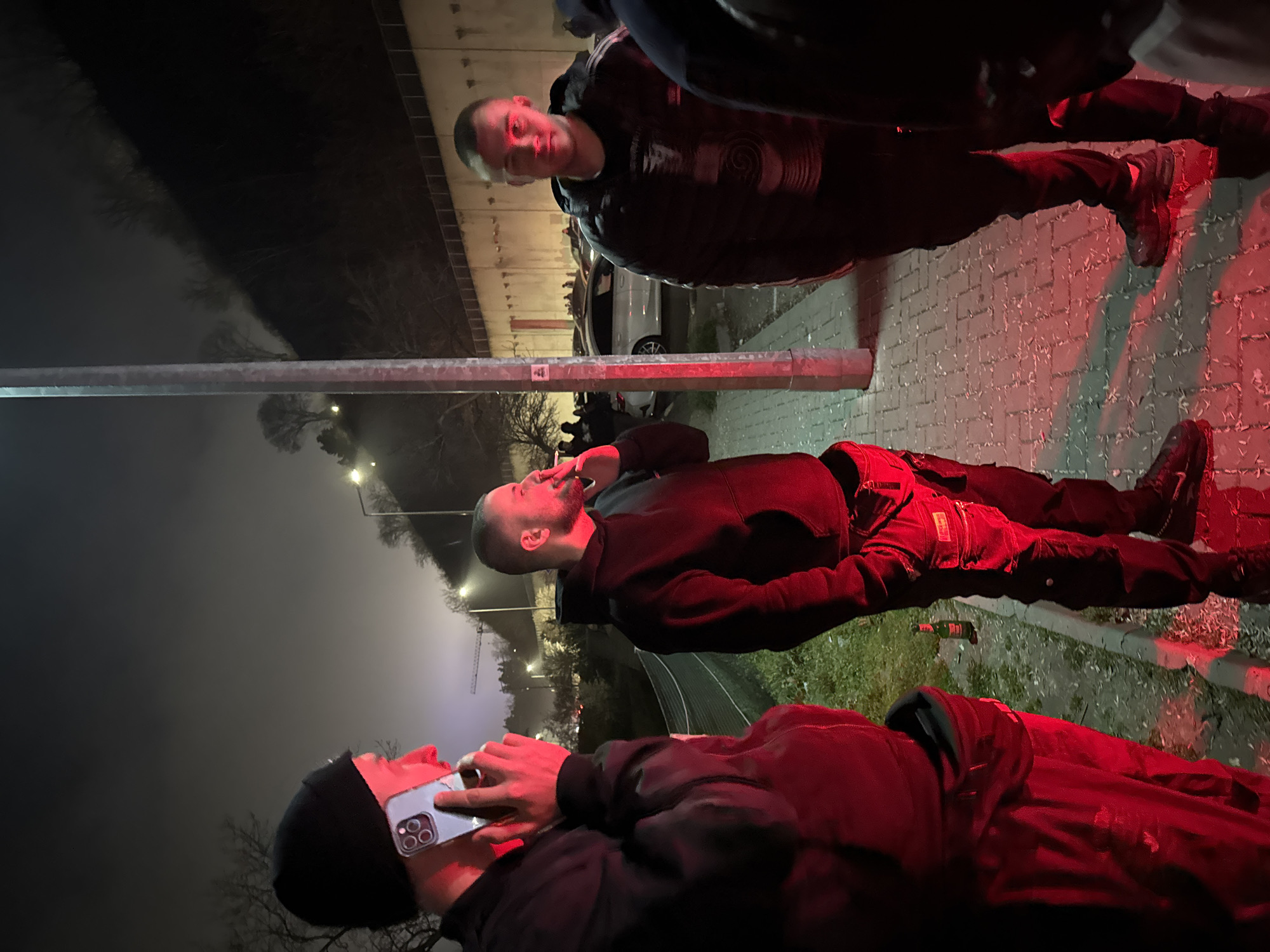 Three males stand on a paved surface at night. One male holds a lit cigarette near his mouth. Another holds a cell phone to his ear. One male looks towards the camera. A metal pole is to the left of the group. In the background are streetlights, trees, and a white vehicle. The group is outdoors. A discarded beer bottle is on the ground. The scene is illuminated with red light, potentially from emergency vehicle lights. Given the streetlights, trees, and a white vehicle in the background, this could be near a road or parking lot. Without more detail, the location of Varna, Bulgaria cannot be confirmed.
