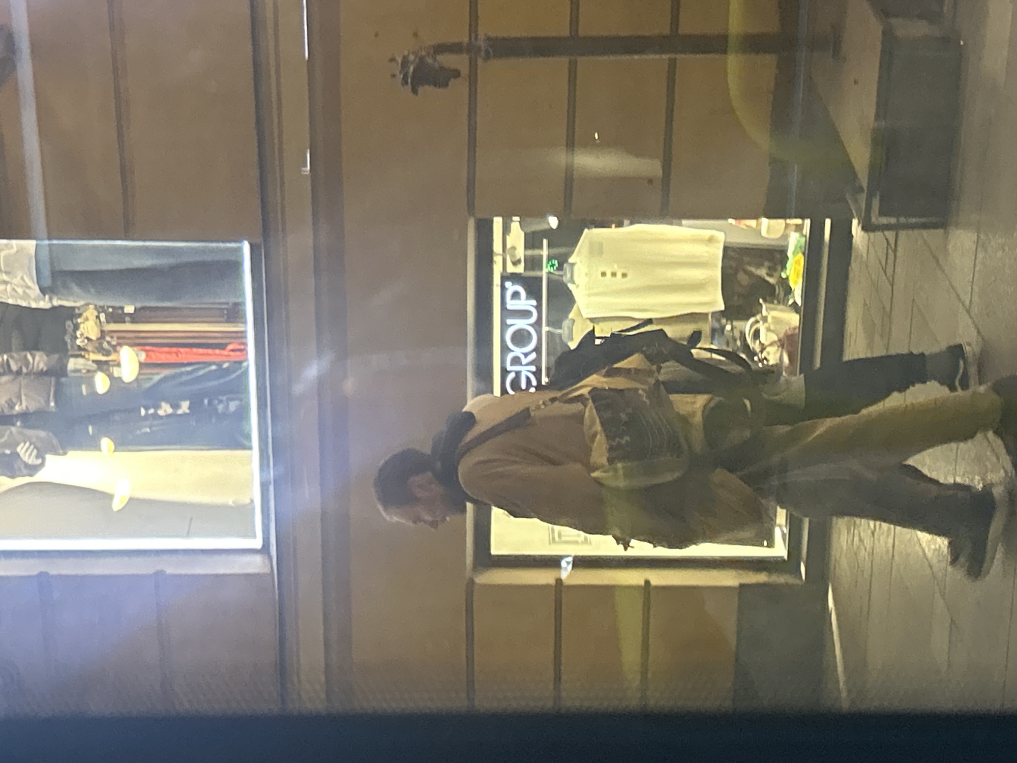 This image captures an urban street scene at night or dusk, likely in a commercial area, given the brightly lit shop windows. The primary subject is a man, viewed in profile, walking past the storefronts. He has a short beard and is wearing a light brown or tan jacket, dark trousers, and dark athletic shoes. A distinctive, patterned shoulder bag is slung across his body.
Behind the man are two shop windows displaying clothing. The window on the left shows what appears to be jeans and other garments on mannequins. The window on the right features a white collared shirt on a mannequin, alongside other clothing items. A sign with the word "GROUP" in white capital letters on a dark background is prominently visible within this shop.
Reflections of the street environment and other people's legs and feet are visible in the shop windows, indicating an active urban setting. The overall lighting suggests it is late evening or night, with artificial light from the shops illuminating the scene. The context provided specifies the location as Saint Petersburg, Russia.