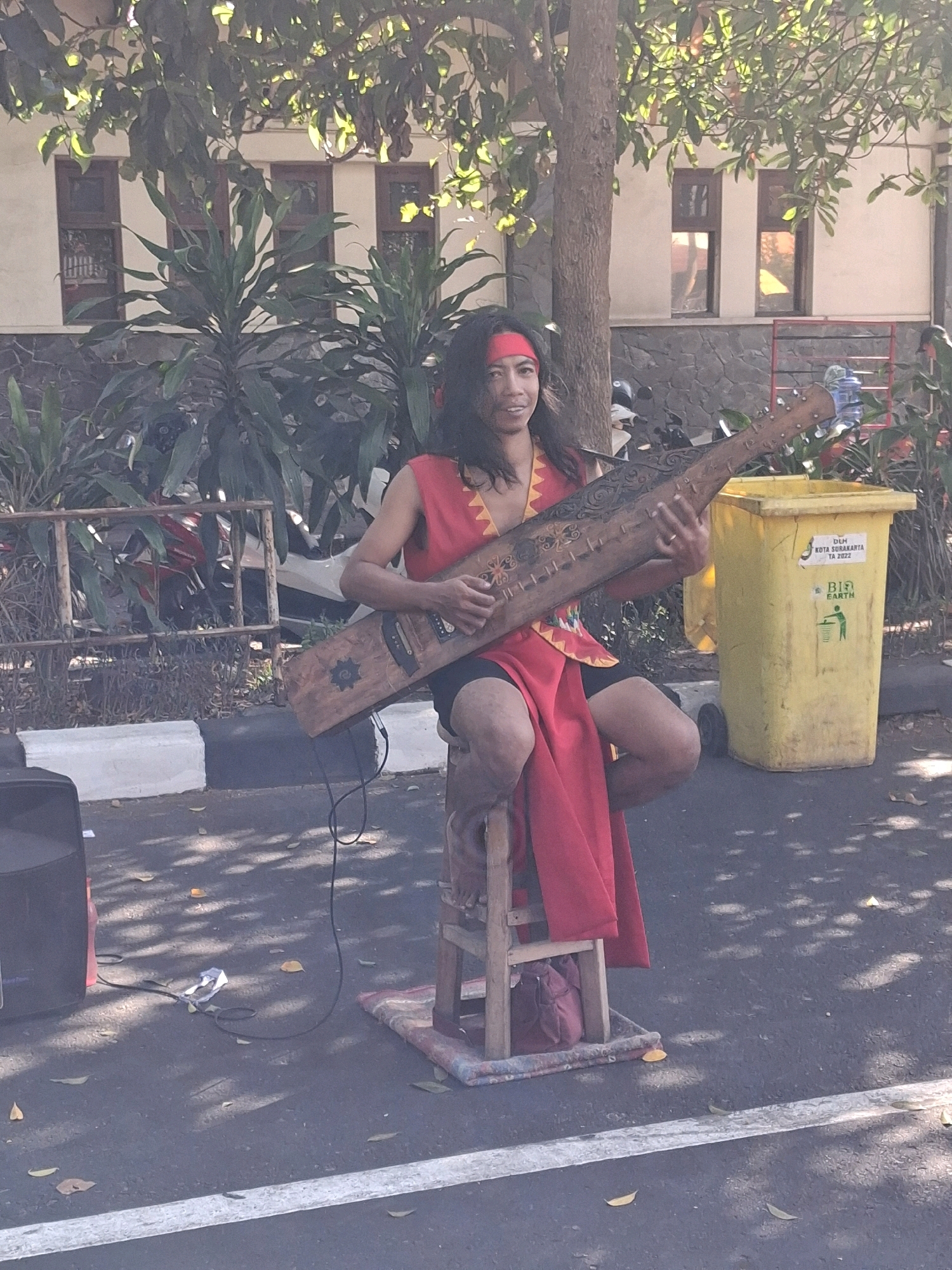 An Indonesian street performer sits on a wooden stool in Surakarta, Indonesia, holding and playing a traditional stringed instrument. The performer, wearing a red sash around their head and a red sleeveless top with yellow trim over black shorts, is seated in front of a building with multiple windows. A large yellow trash receptacle with text in Indonesian is visible to the right of the performer. To the left, a black speaker sits on the asphalt ground, with cables extending towards the performer. The ground is paved and marked with a white line. Trees with dappled sunlight filtering through their leaves cast shadows on the scene. The background shows a fence and parked motorcycles.