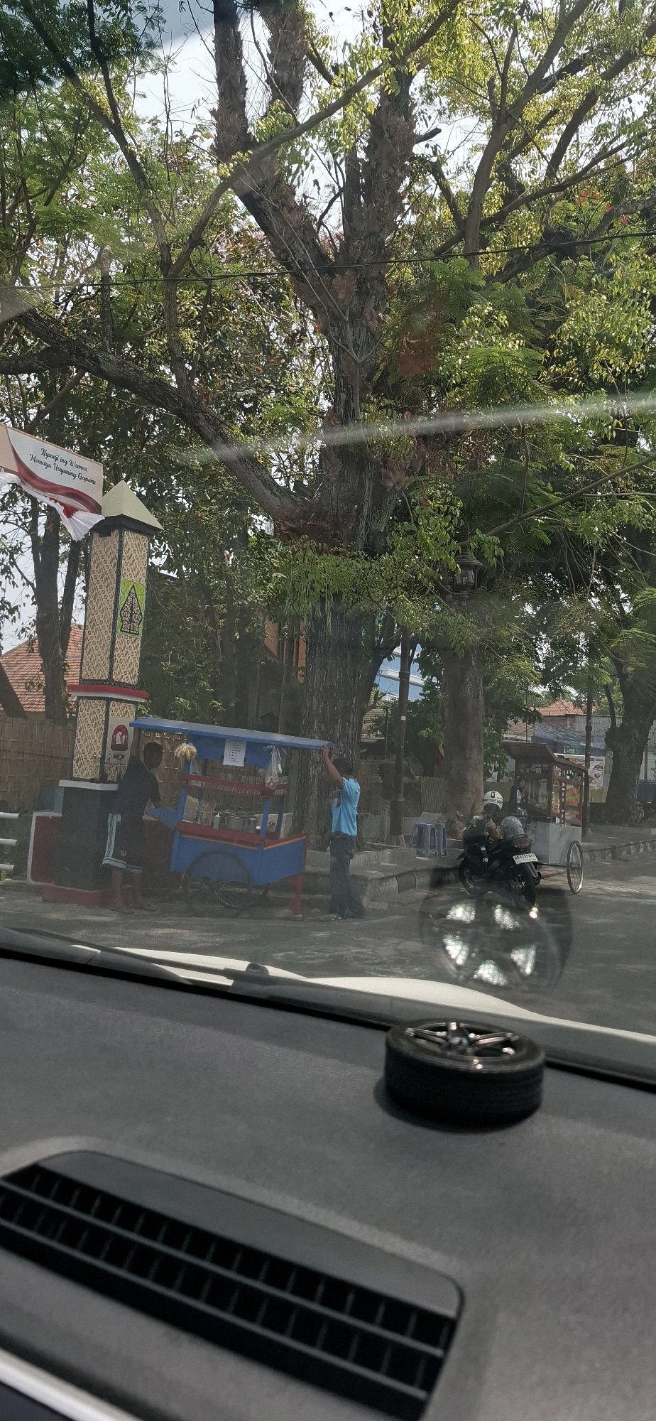 In Surakarta, Indonesia, an outdoor scene is captured from inside a vehicle. Dominating the background is a large, old tree with extensive branches and foliage, partially obscuring the sky. In the mid-ground, a colorful food cart, painted blue and red, is positioned near a decorative pillar adorned with cultural motifs and a banner. Two individuals are near the cart, one appearing to adjust its canopy. Several motorcycles are parked to the right of the cart, with one rider visible. The foreground shows the interior of the vehicle, with the dashboard and a small decorative tire visible. The environment suggests a bright, daytime setting.