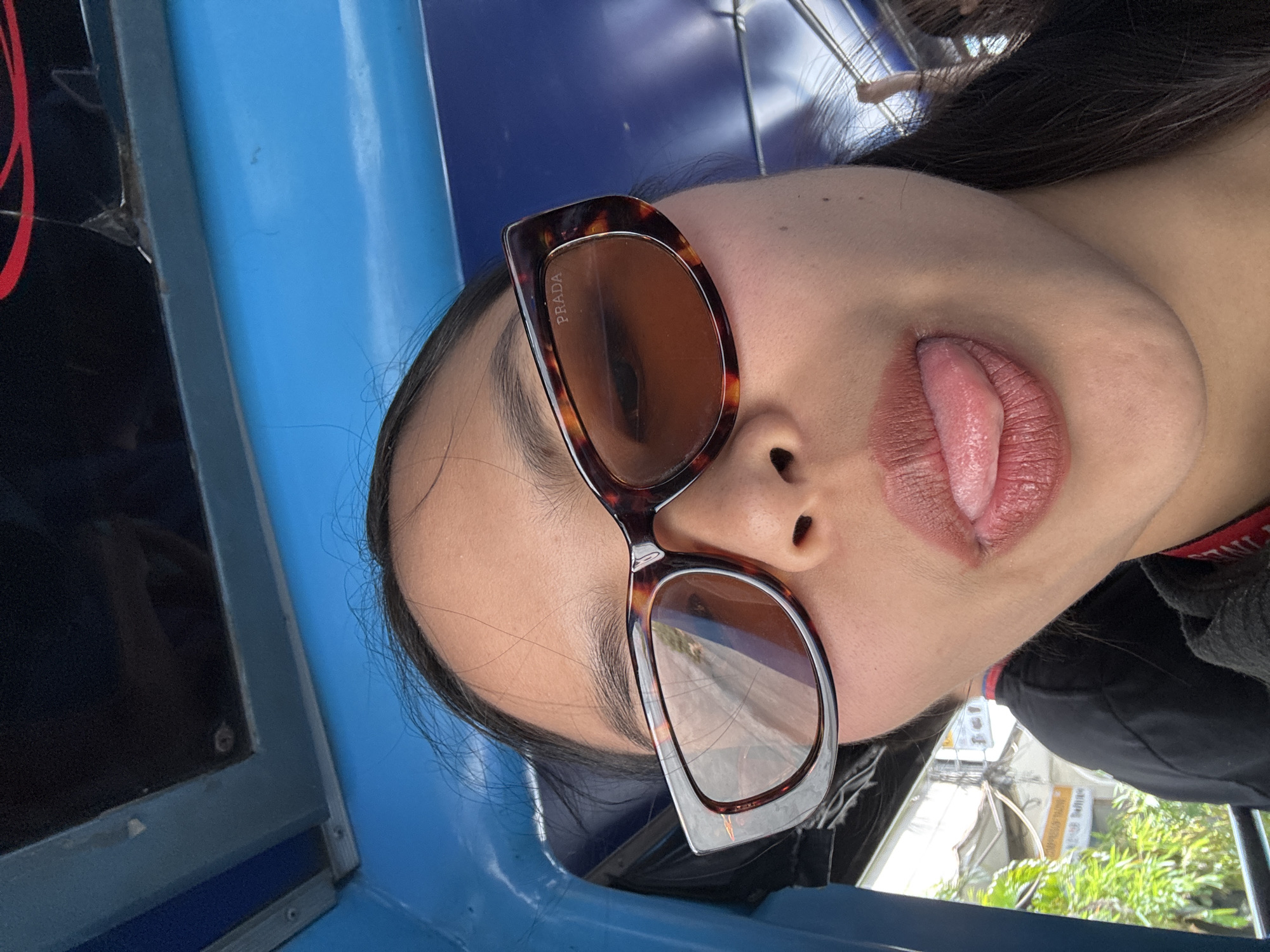 A person, likely female, is pictured from a low angle, wearing sunglasses with the brand name "PRADA" visible on the lens. The person's tongue is protruding from their mouth. The background indicates the person is riding in a blue vehicle, possibly a tuk-tuk or similar open-air transportation, with signs of urban scenery, including vegetation and buildings, suggesting a city environment. The specific location is indicated as Davao City, Philippines.