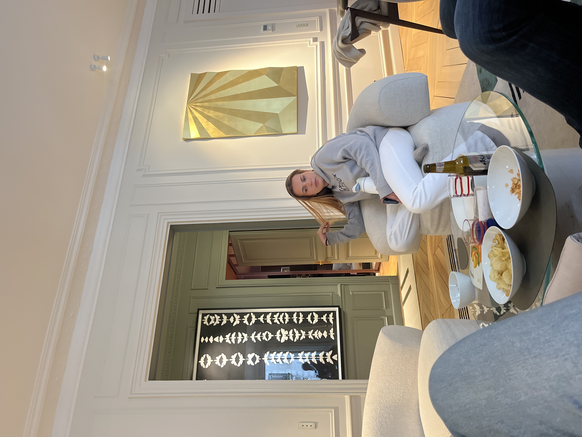 The interior is that of an apartment in Paris, France. The room features white walls with ornate, decorative molding. On the wall, a modern, gold-colored geometric artwork is hung. Below that, a black framed picture is visible through an open doorway.
A woman in a grey hoodie and white pants is lounging on a light-colored, modern-style sofa. She is looking at the camera.
A glass-topped coffee table is in front of the sofa. On the table, there's an opened bottle of wine, glasses, and two bowls filled with snacks, appearing to be chips and nuts. At least two other individuals can be seen partially in the shot, presumably sitting on the sofa.