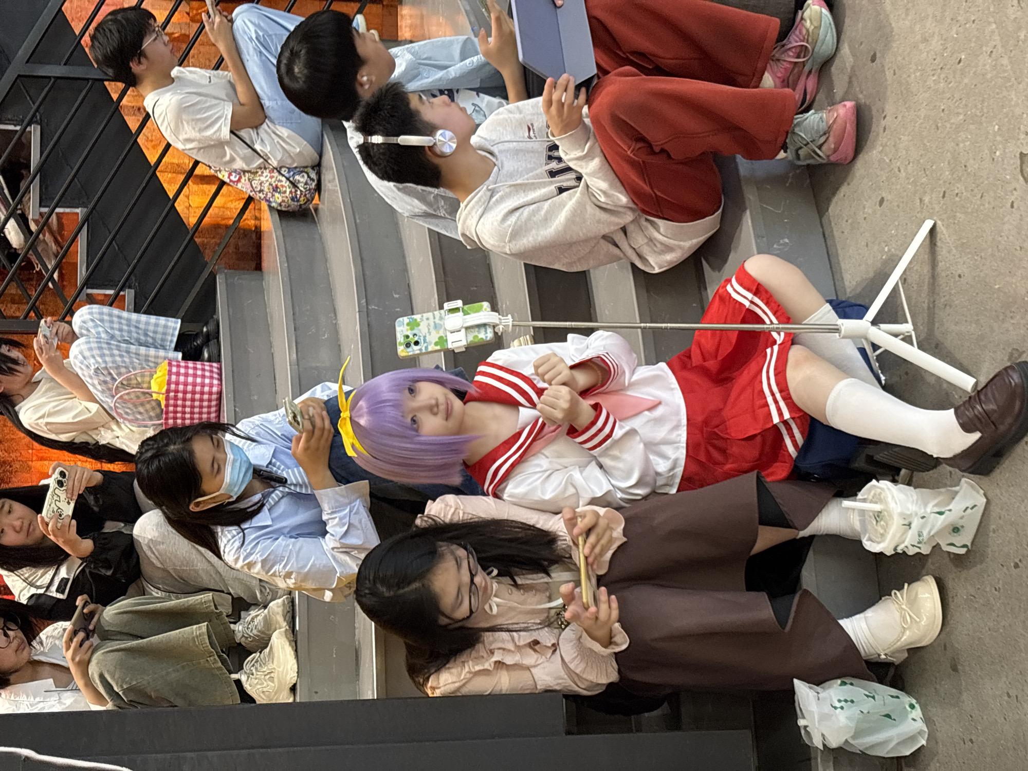 A group of young people are sitting on outdoor steps. Several are using phones. One young woman with purple hair and a red and white sailor-style outfit has a phone mounted on a selfie stick. The steps are near an orange building with dark metal railings. Cups in plastic bags are next to the feet of a seated person. The image was taken in Liangjiang, China.