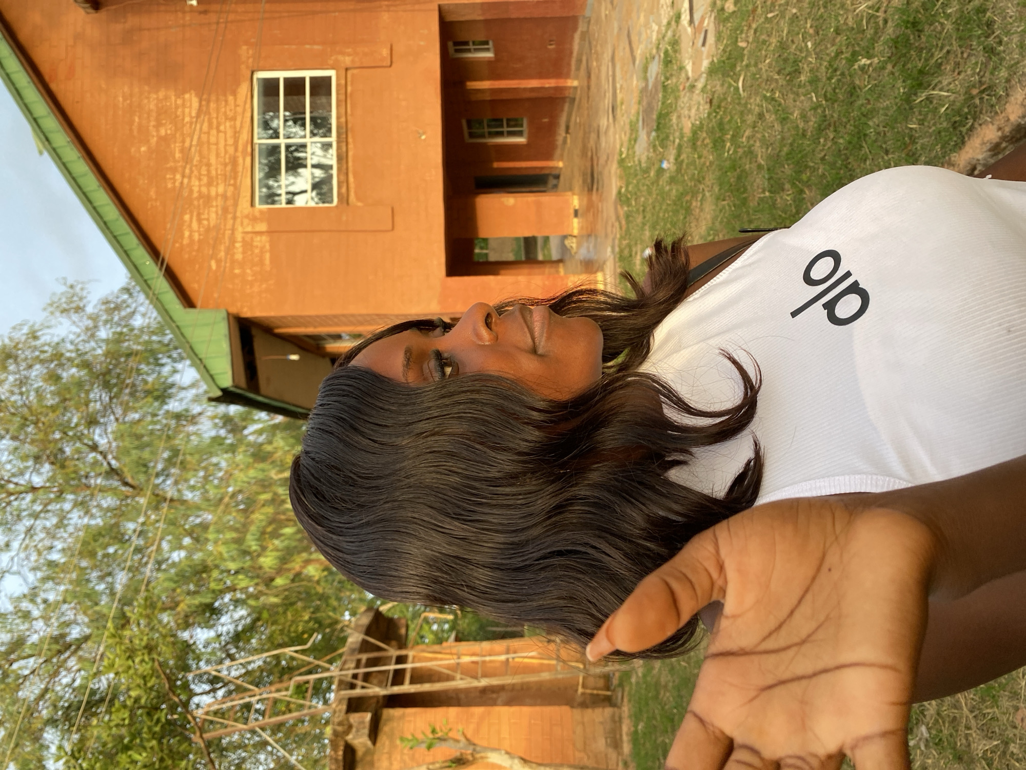 A woman wearing a white shirt with the word "alo" on it is lying on the grass in front of a brick building with a green roof, likely in the city of Unknown, Nigeria. The building has windows and a columned walkway. Trees and other greenery are visible in the background. The woman is looking up with a slight smile, and her hand is outstretched towards the camera. The image is slightly overexposed and has been rotated.