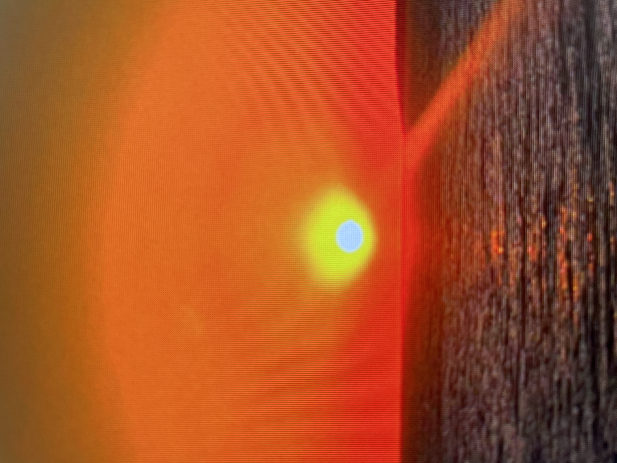 The image appears to be a close-up photograph, possibly taken with a digital device or a still from a video recording, given the horizontal scan lines visible across the frame.
On the left side, there is a predominantly orange and reddish-orange color field. Within this area, a lighter, yellow-to-white spherical shape with a slightly bluish center is visible. The surrounding hues blend into the orange background, creating a diffuse gradient effect.
To the right, a textured, dark-colored surface abruptly begins, indicating a sharp transition. This surface has a vertical grain-like texture with hints of orange and brown interwoven within the darker shades.
The image does not provide any visible information regarding a specific location or subject, making it impossible to definitively ascertain if the scene is located in the city of Zlatograd, Bulgaria. There are no discernible people, actions, or interactions within the frame.