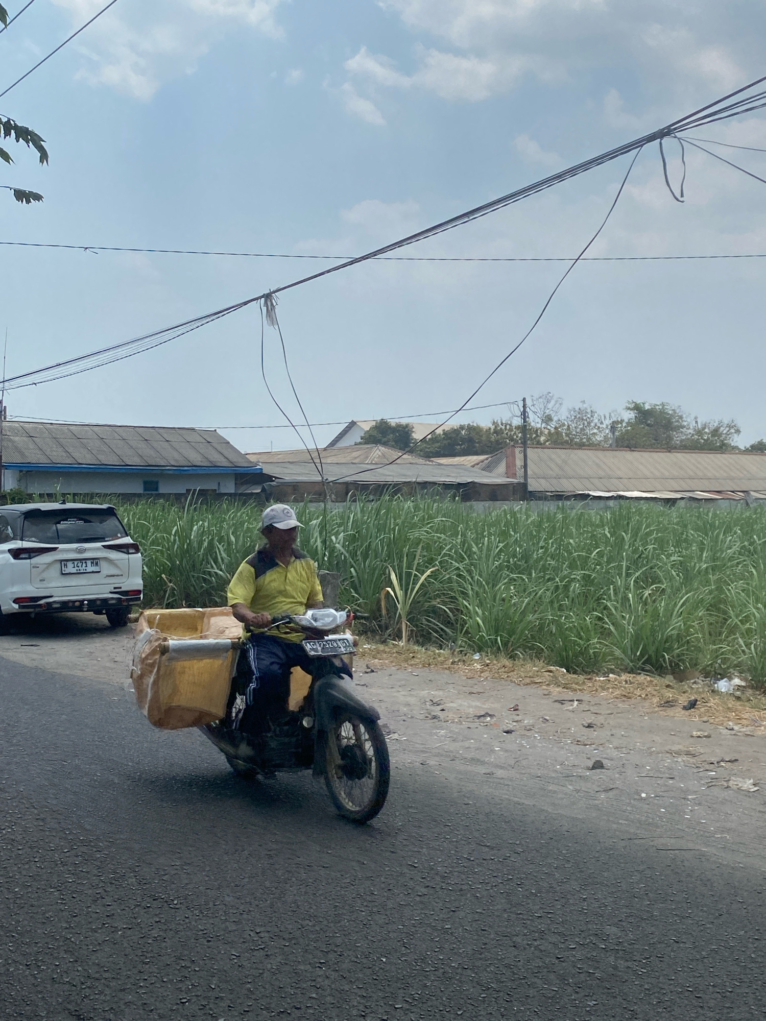 A man wearing a yellow shirt and a white cap rides a motorcycle loaded with yellow containers. He is traveling on a paved road in front of a field of tall green grass, likely sugarcane. In the background, there are buildings with corrugated metal roofs. A white SUV is parked on the side of the road. The scene appears to be in the city of Kediri, Indonesia, given the Indonesian license plates on the vehicles. The sky is partly cloudy.